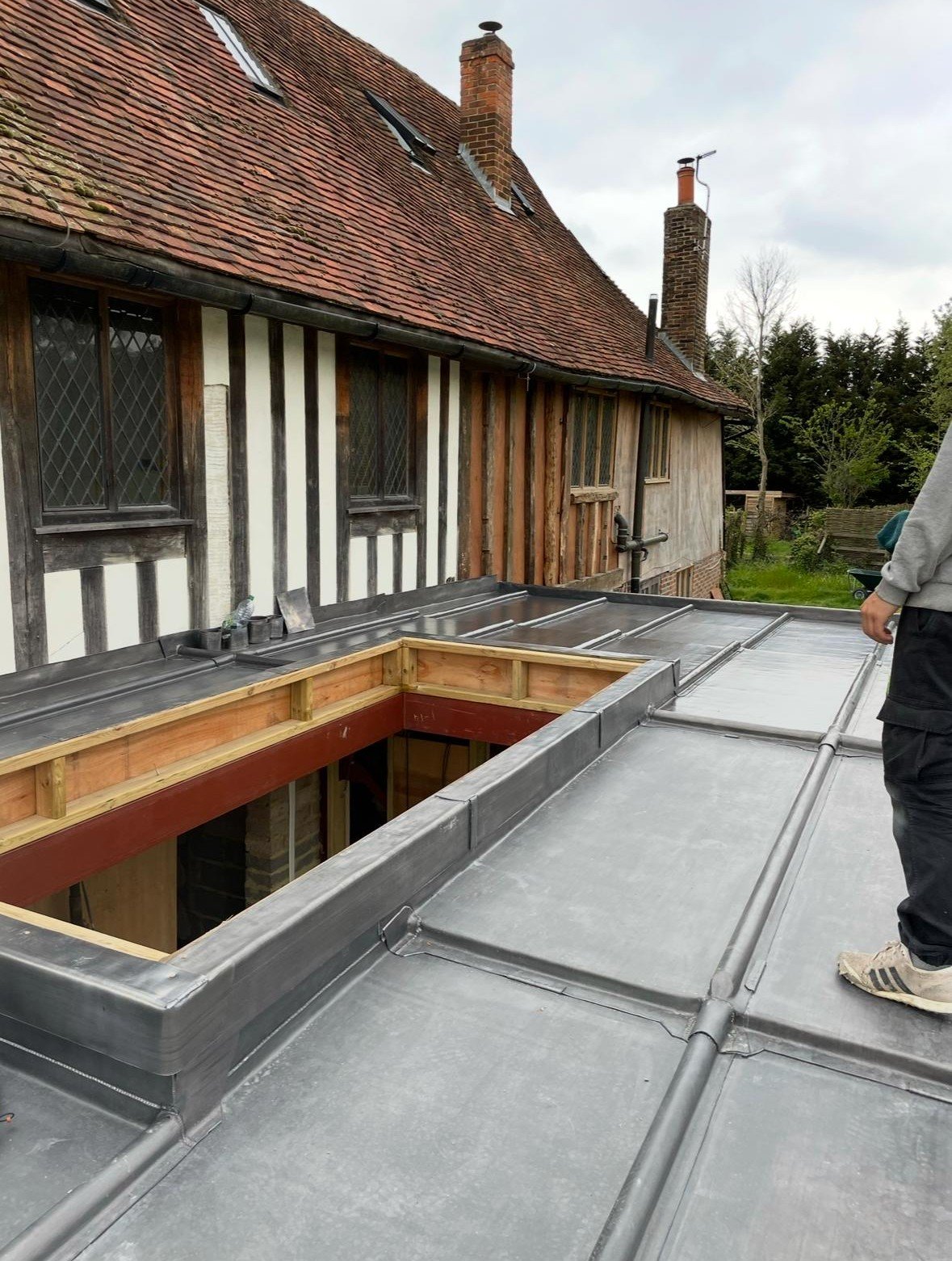 Rooftop construction with metal flashing and wooden framing, adjacent to a traditional house with timber framing and a red tiled roof.