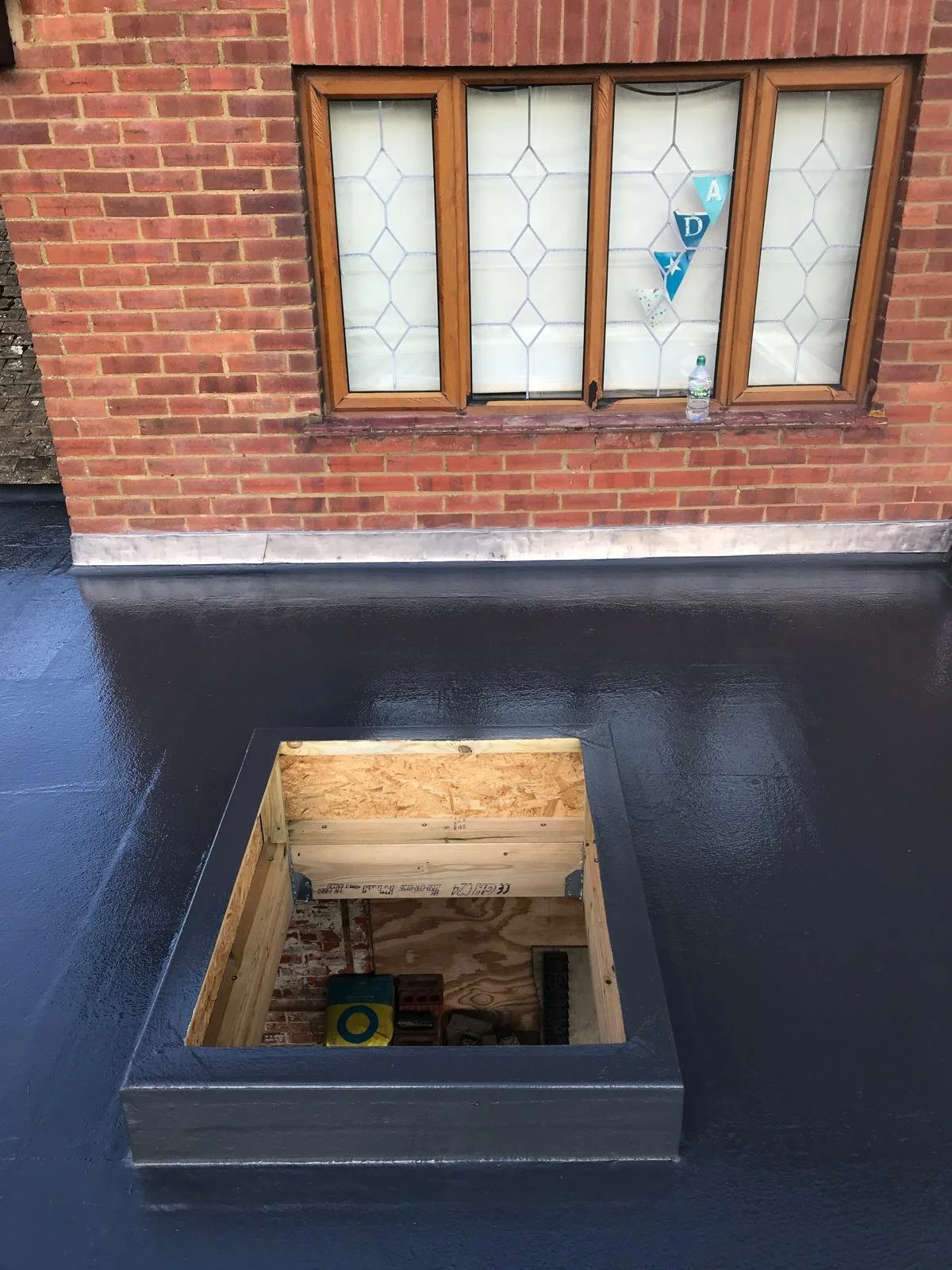 View of a brick building with a wooden-framed window, decorated with blue A, D, and Y banners, and a water bottle on the window sill. The photo is taken from a rooftop, showing an open square hole with a black waterproof coating around it, revealing 