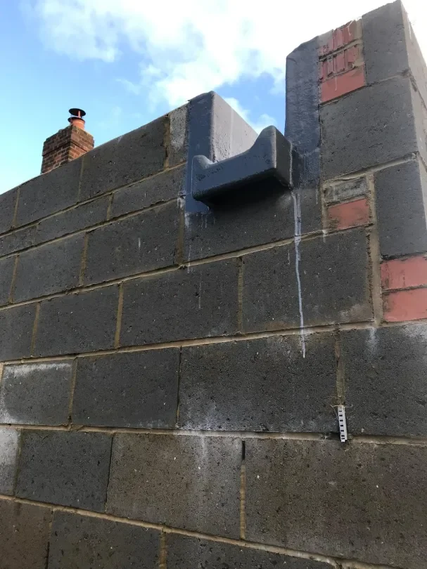 Close-up of a brick wall with a metal object attached to the top corner, possibly a corner guard, with water leaking from it. Part of a chimney is visible in the background against a partly cloudy sky.