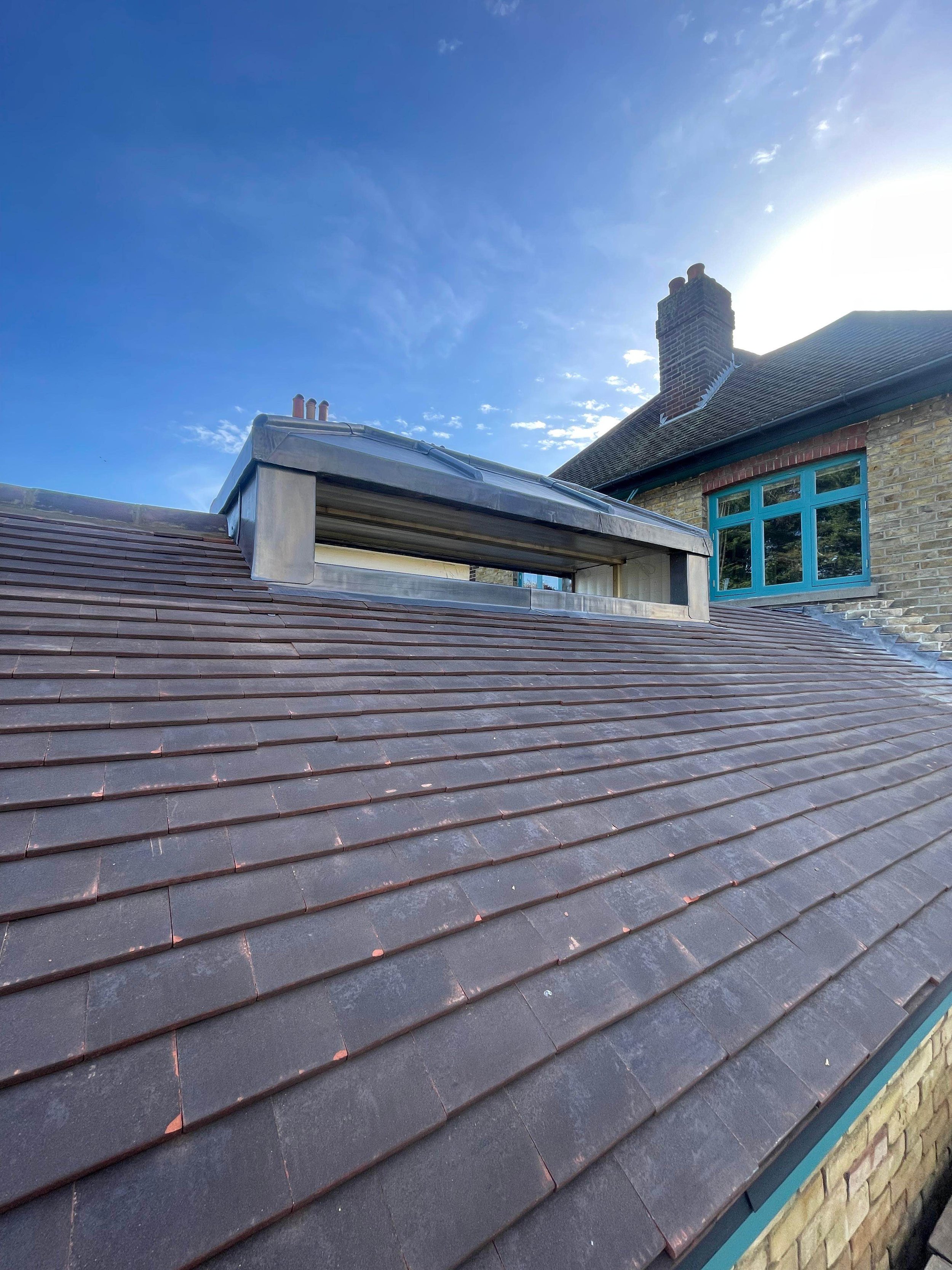 Close-up of a pitched roof with brown tiles, featuring a skylight window and a brick house with blue window frames under a clear blue sky.