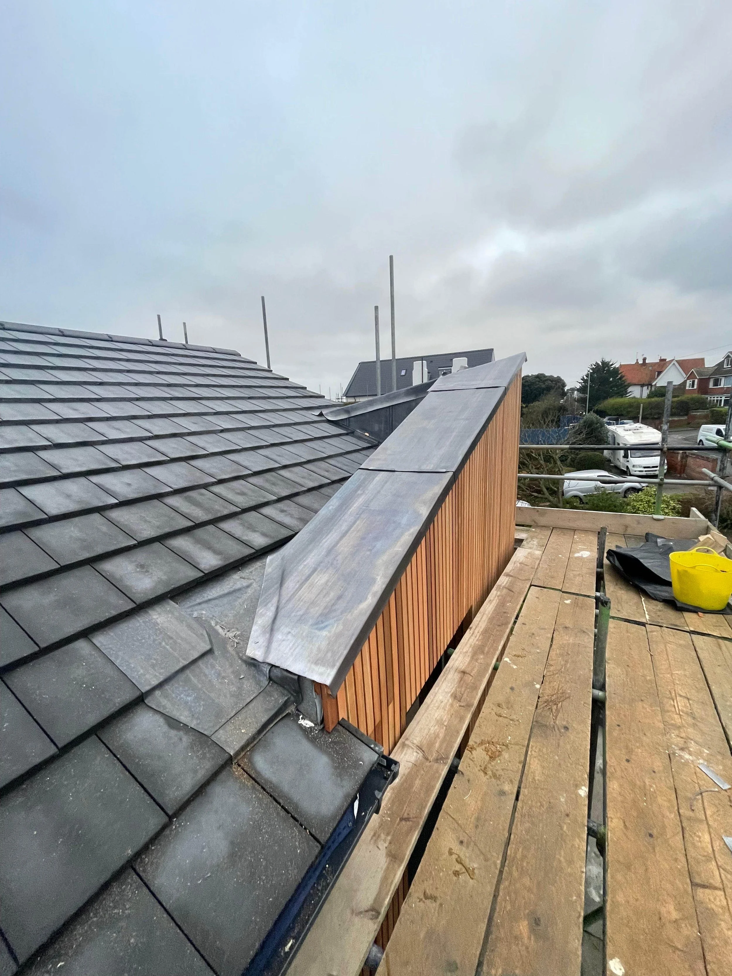 Roof construction scene with tiles, a wooden wall, and metal flashing on a cloudy day.
