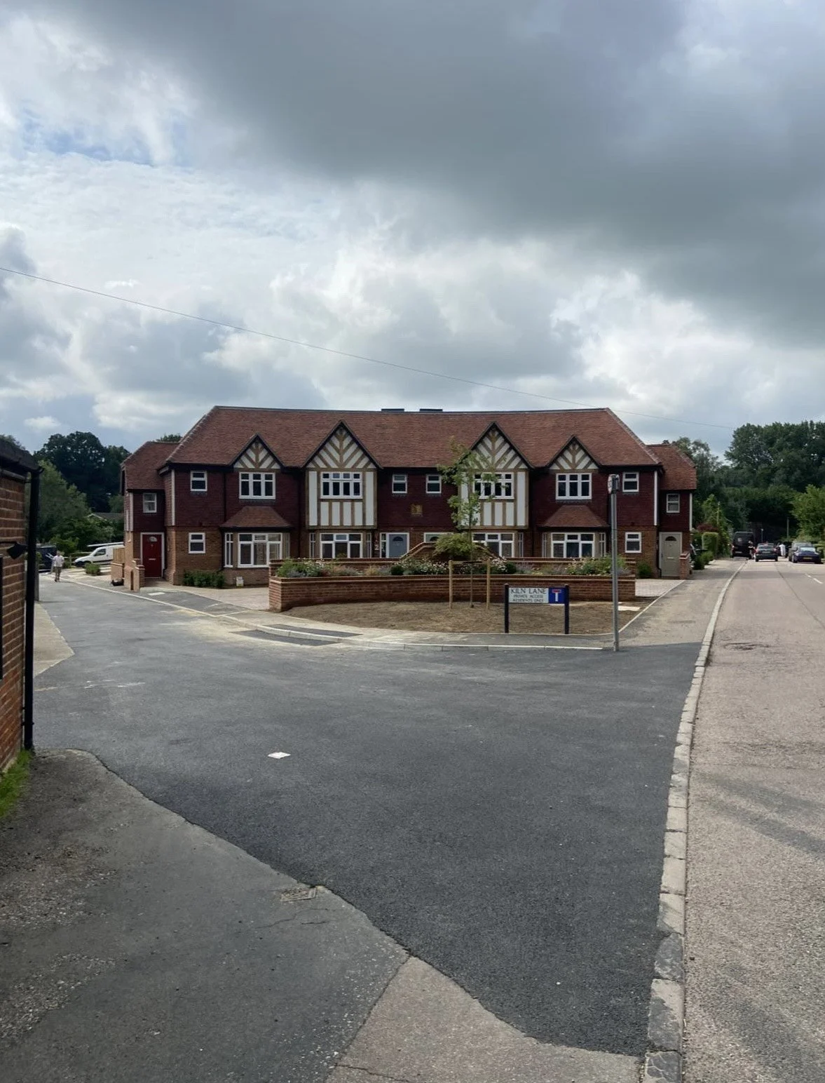 A red brick residential building with multiple gabled roofs and white-framed windows, surrounded by a small brick wall and parking spaces, under a cloudy sky.