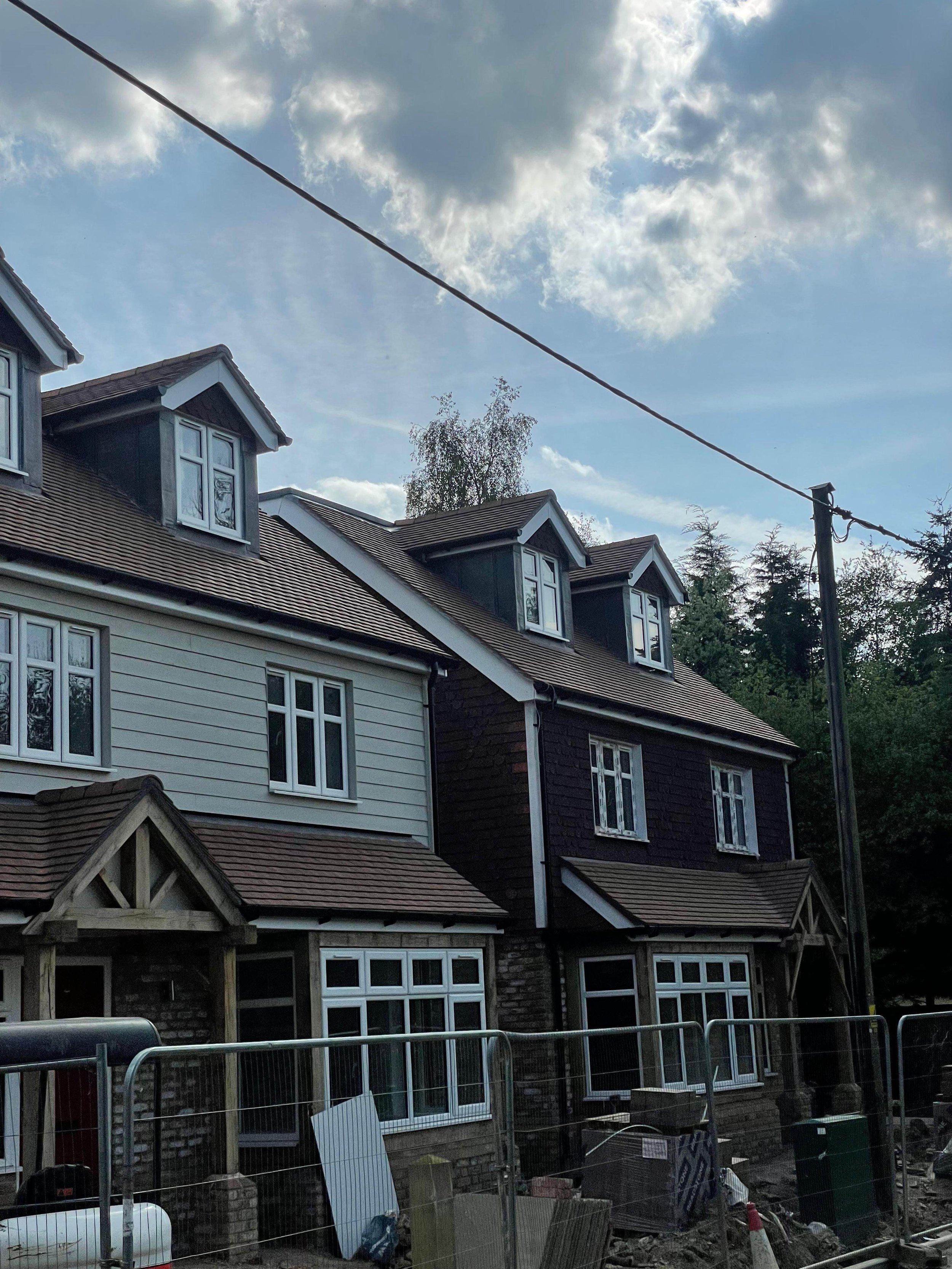 Side view of a multi-story house with white and dark brown siding, multiple bay windows, and dormer windows on the roof. Construction fencing and supplies in the foreground, cloudy sky above.