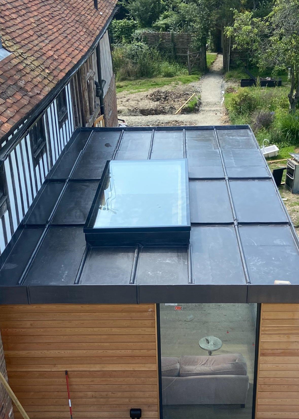A building with a metal roof and a large skylight, with a view of a backyard with a dirt path, trees, and a wooden fence.