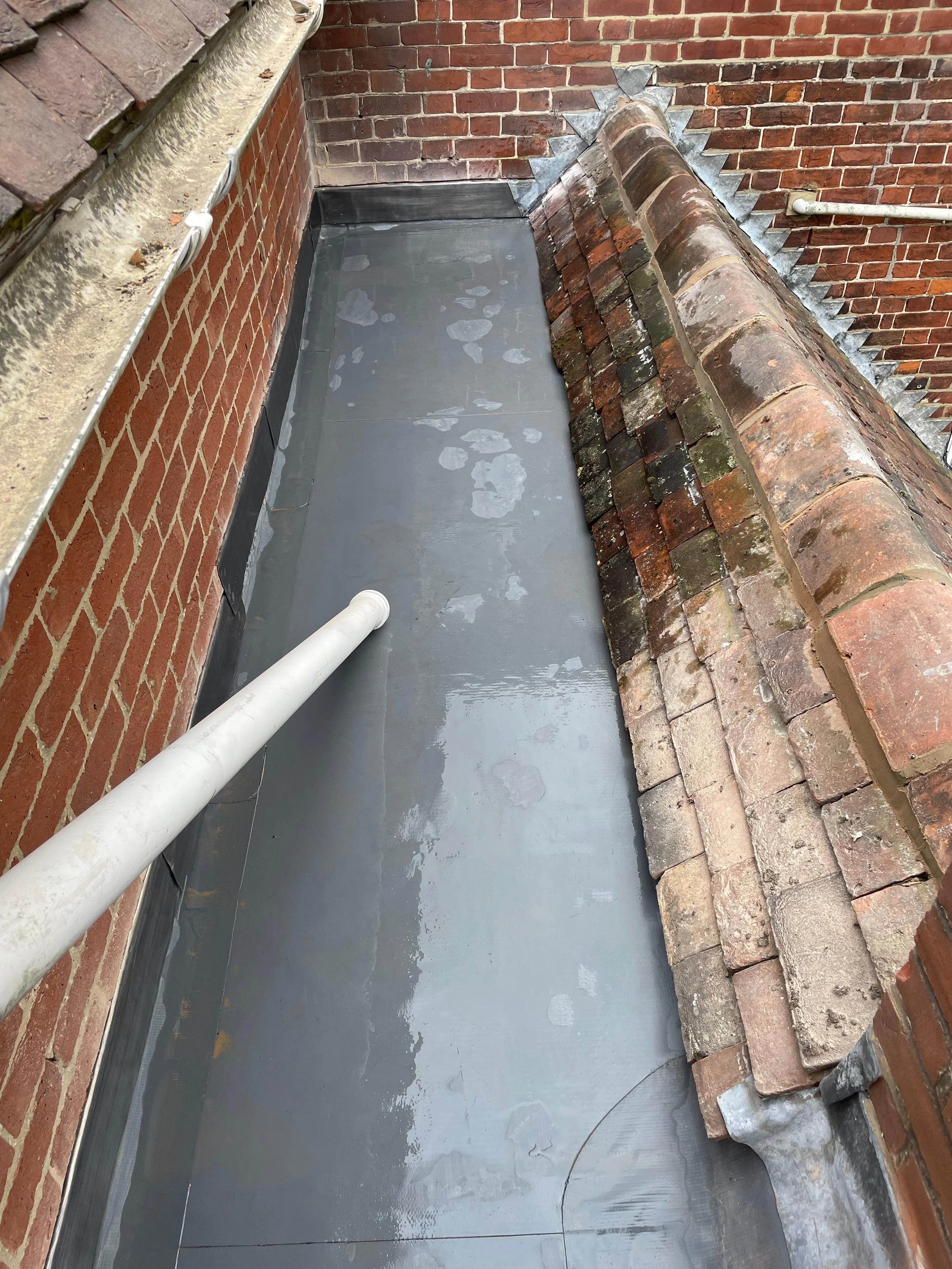 View of a flat roof area with a gray waterproof membrane, brick walls, and a drain pipe. The roof is bordered by old, weathered roof tiles and brick walls.