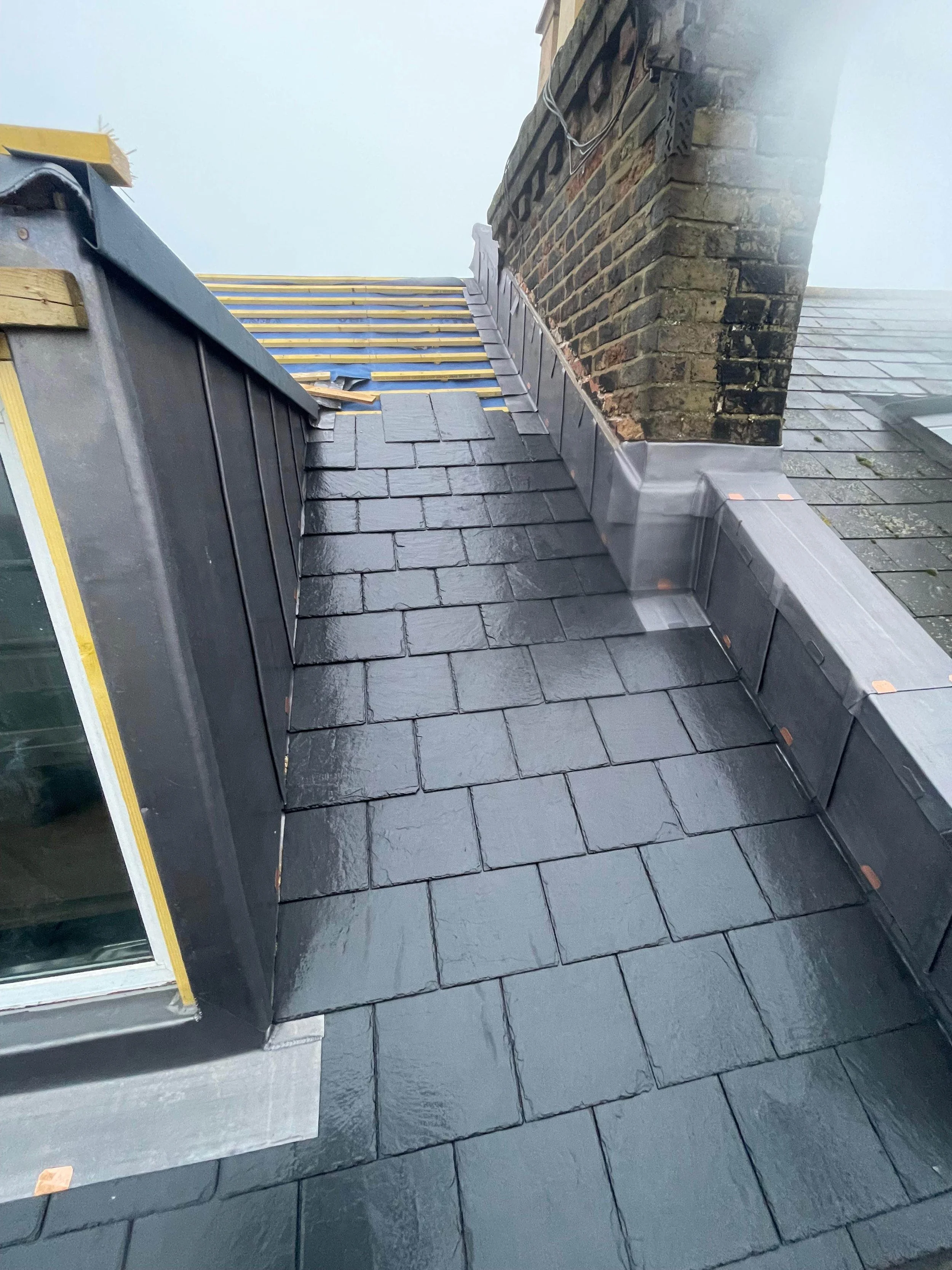 Roof with black slate shingles and a chimney, showing detailed roofing work including flashing around the chimney and partial installation of new shingles.