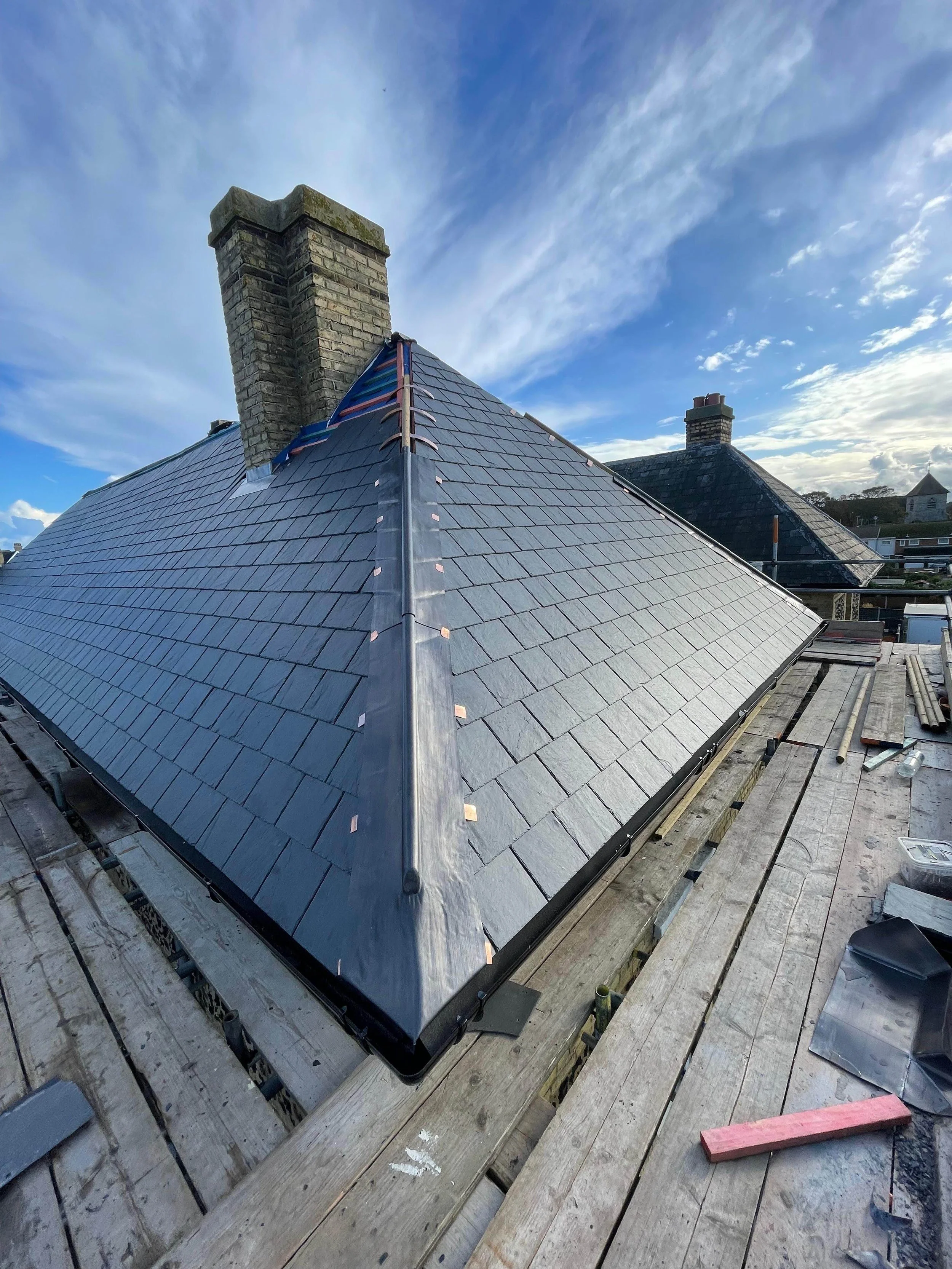 A roof with new slate tiles under construction, scaffolding, brick chimneys, and a sky with clouds.