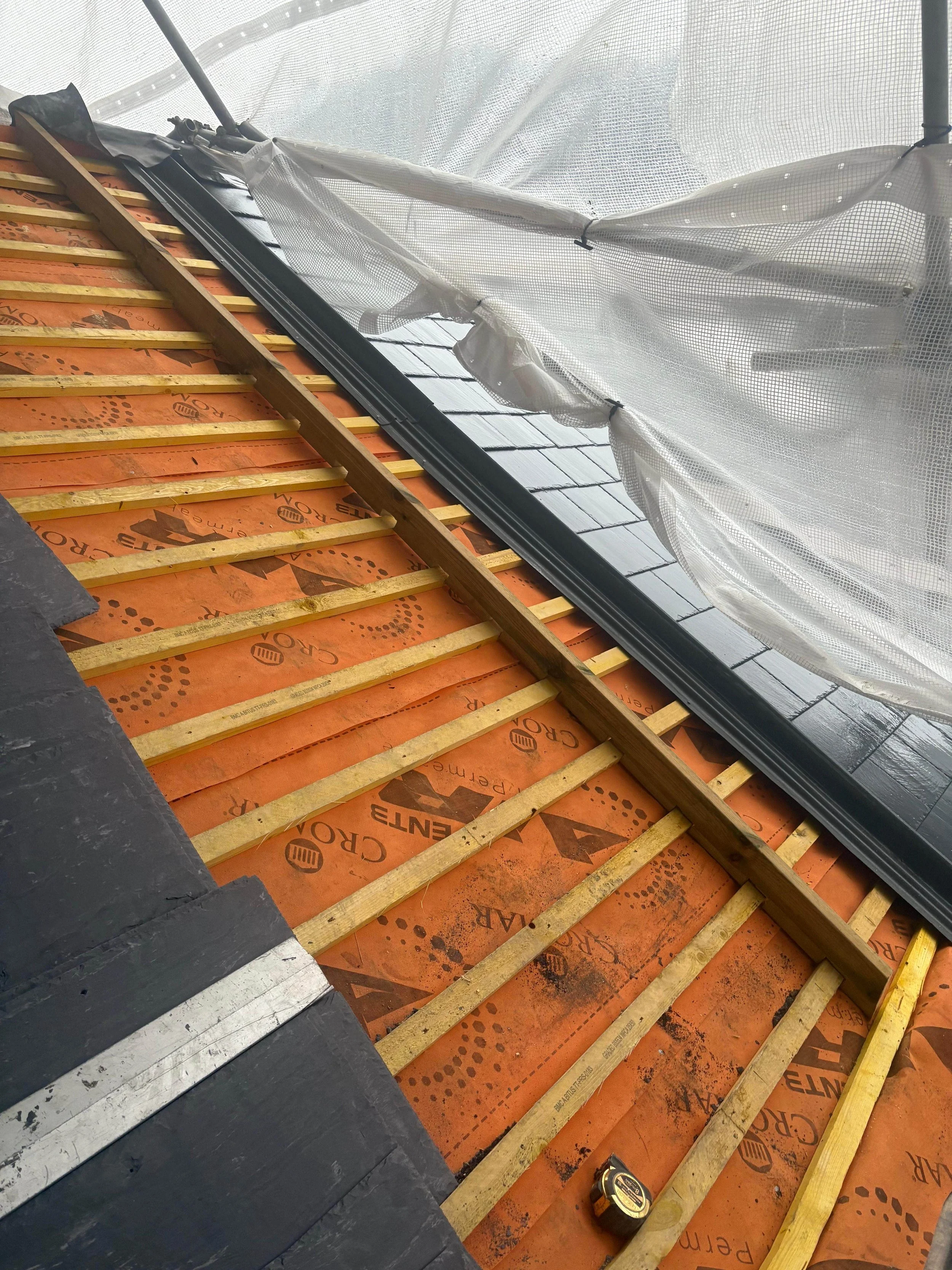 Roof under construction with black shingles, wooden battens, orange roofing underlay, and a partly installed black metal flashing, surrounded by scaffolding and protective sheeting.