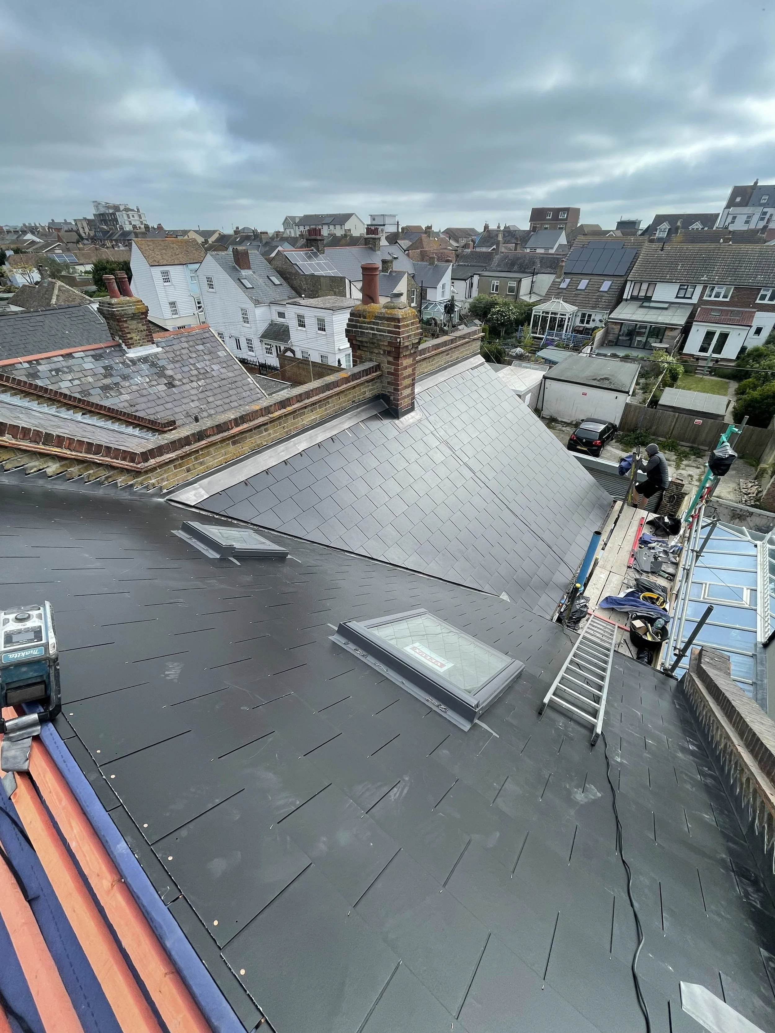 View of rooftops with two skylights, construction scaffolding, and workers on a gray slate roof in a residential neighborhood under cloudy sky.