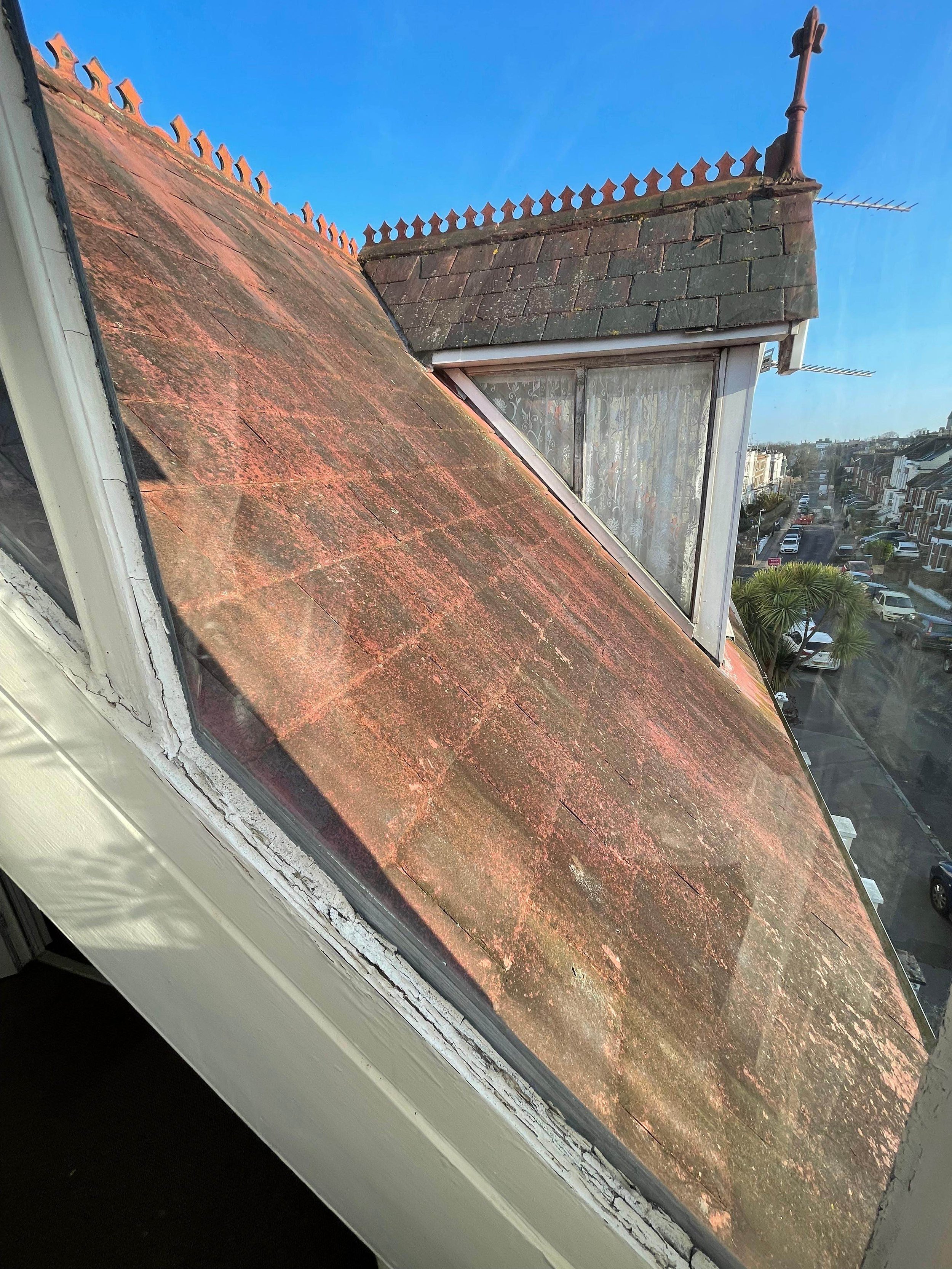 View of rooftops and a street from a window, showing weathered, moss-covered brick roofs with a clear blue sky.