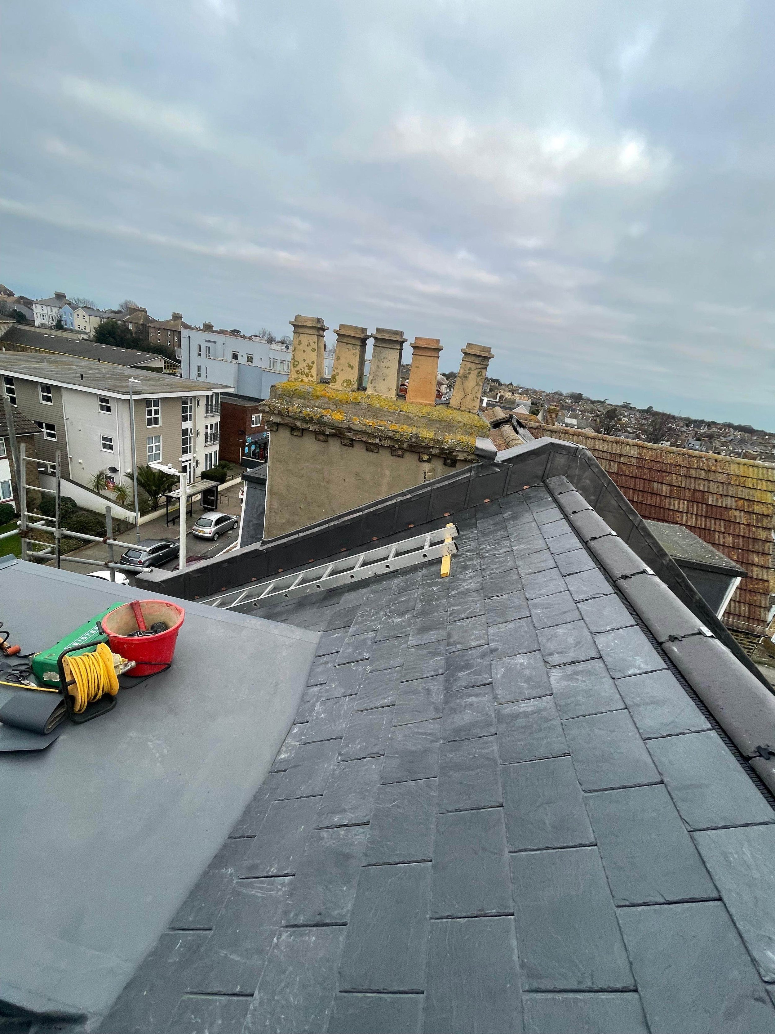 View of a residential rooftop with tools and equipment, including a red bucket, yellow extension cord, and green toolbox, overlooking neighboring buildings and a cloudy sky.