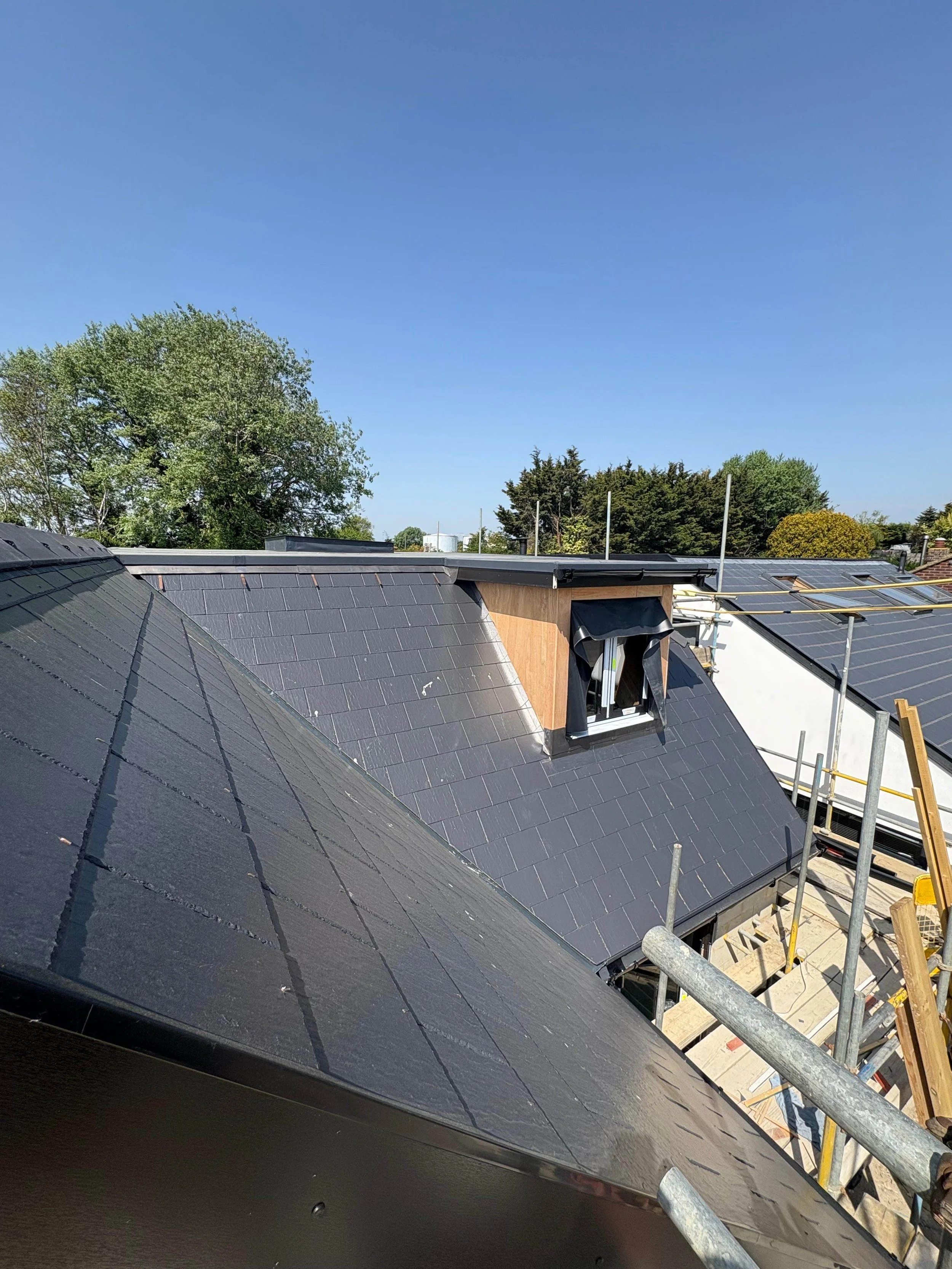 View of a roof with black tiles, a small dormer window with a black frame and a black awning, and construction scaffolding on the side under a clear blue sky with trees in the background.
