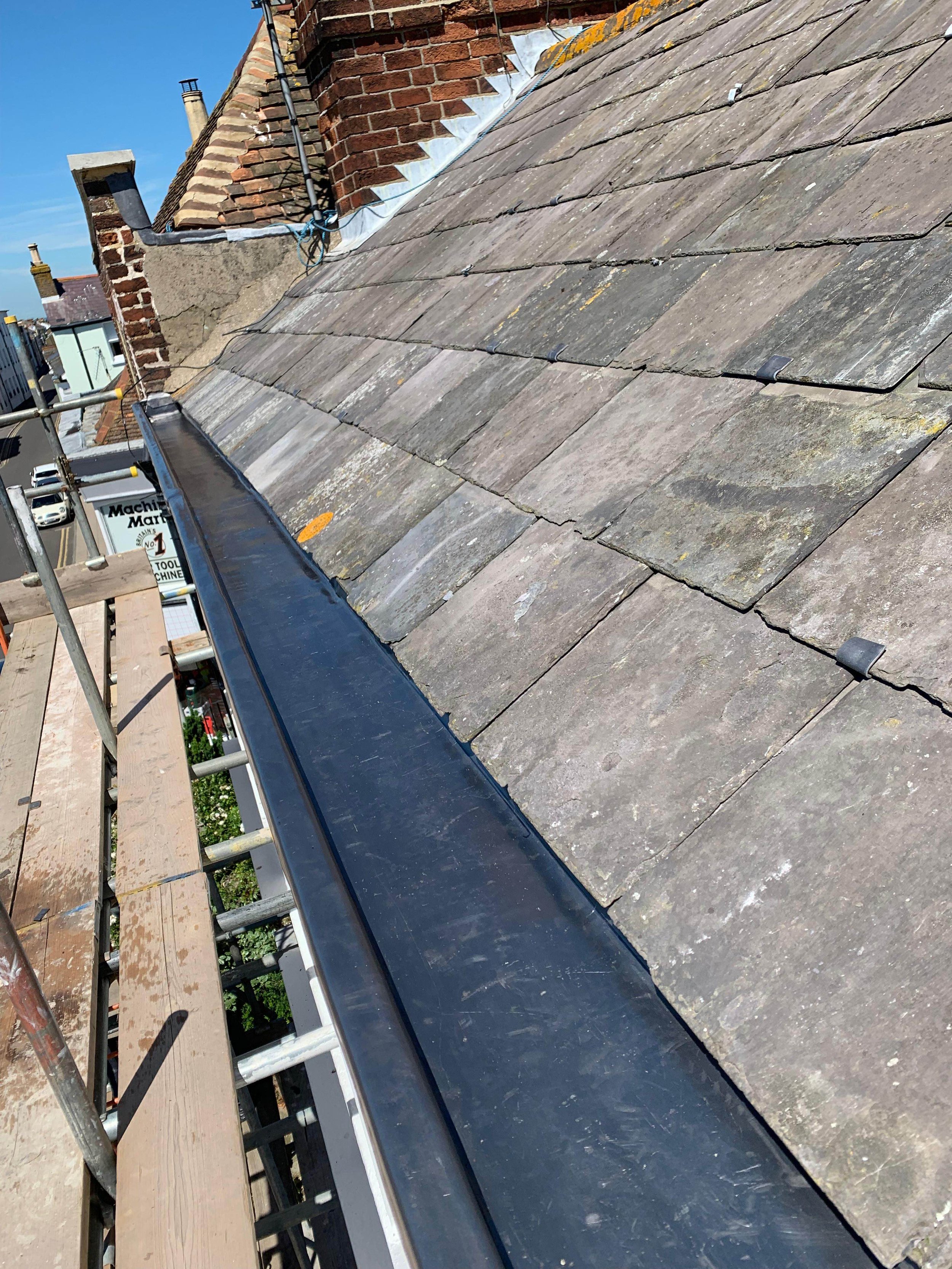 Close-up view of a sloped roof with weathered grey tiles and a black metal gutter along the edge, scaffolding and buildings in the background.