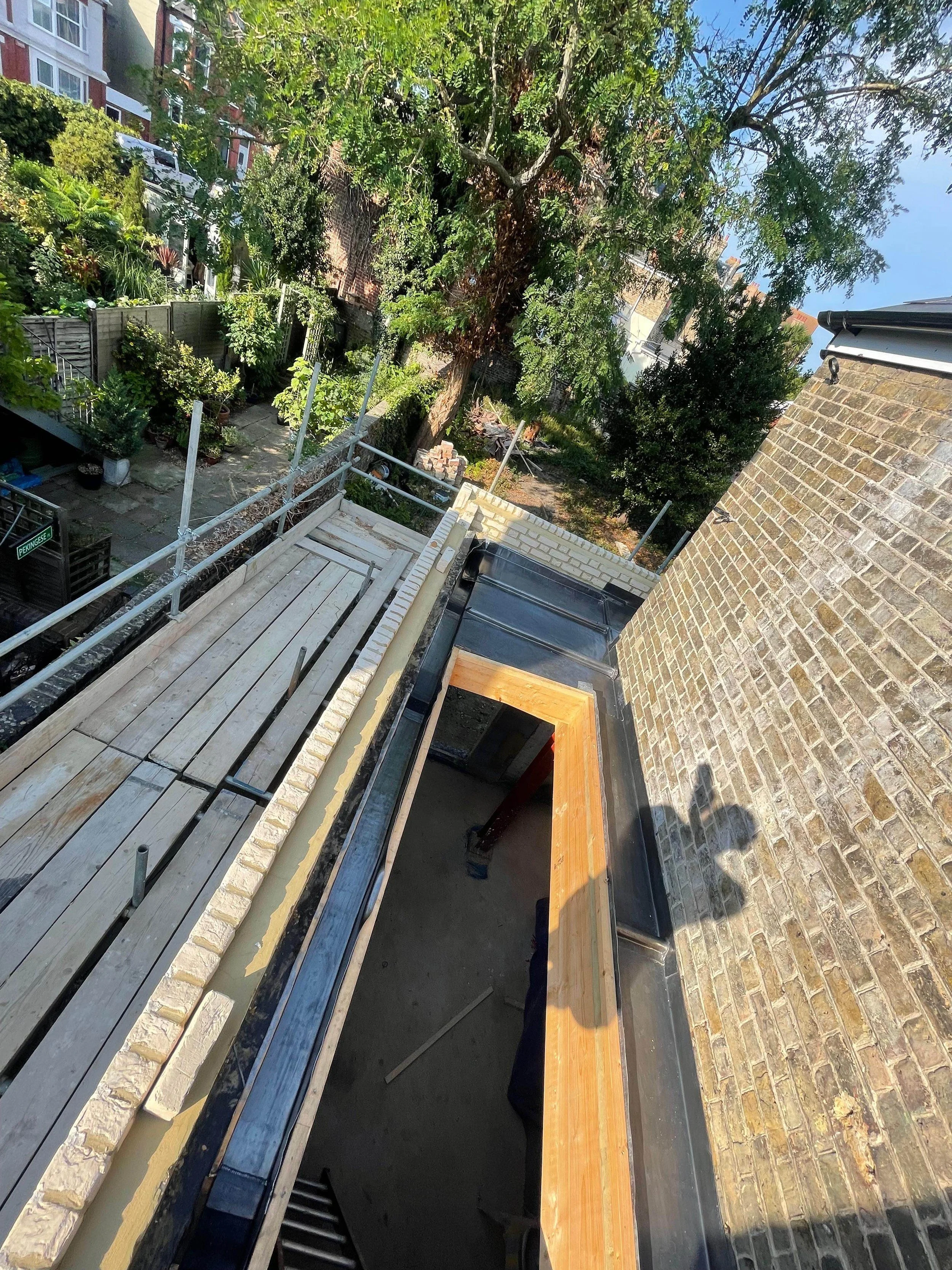 View from a building showing a construction project on a roof with scaffolding, wooden framing, a brick wall, and a lush green backyard with trees and plants.
