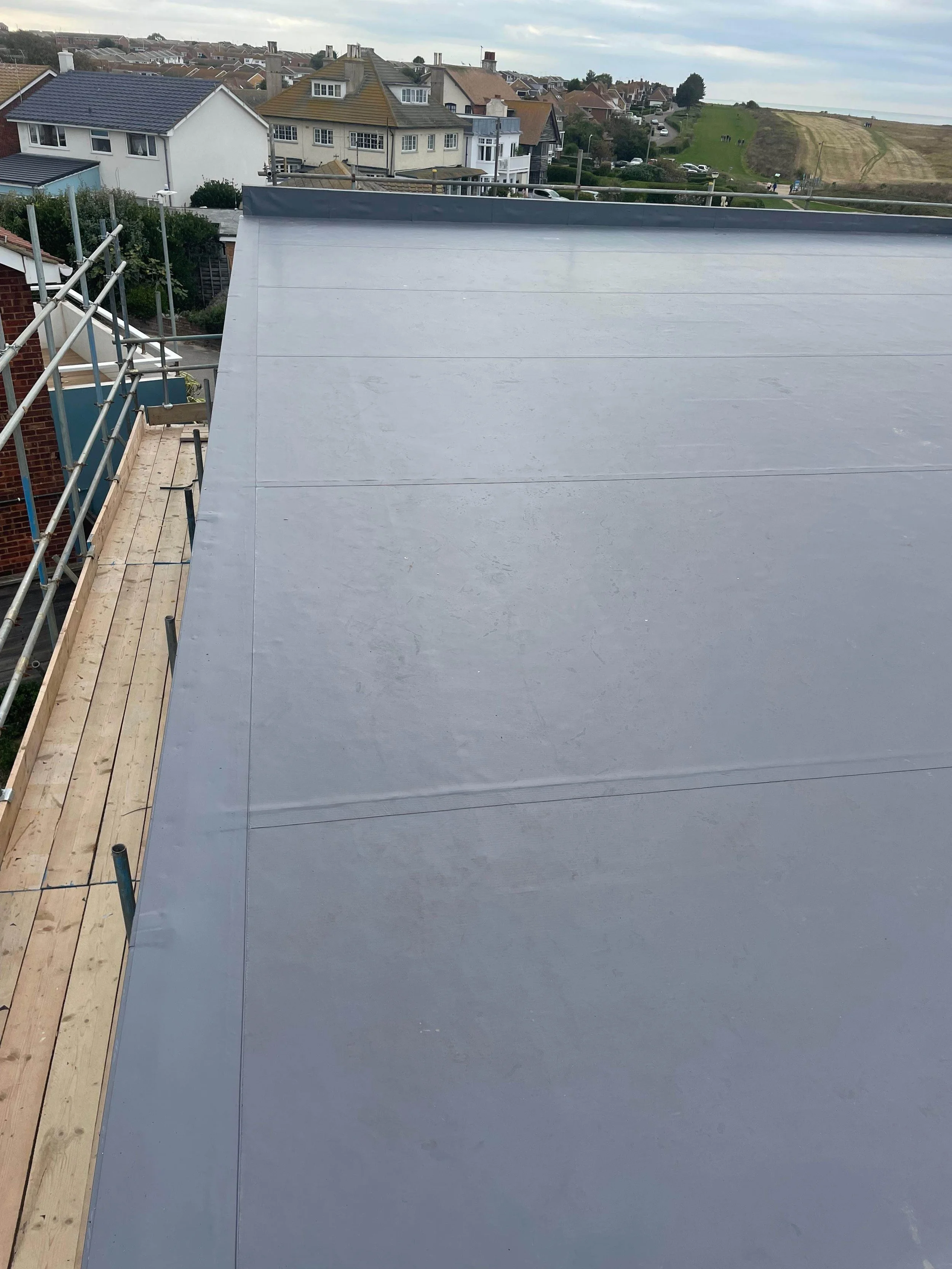 Photo of a large flat roof with a smooth grey surface, part of a building under construction, with houses and a grassy hillside in the background.