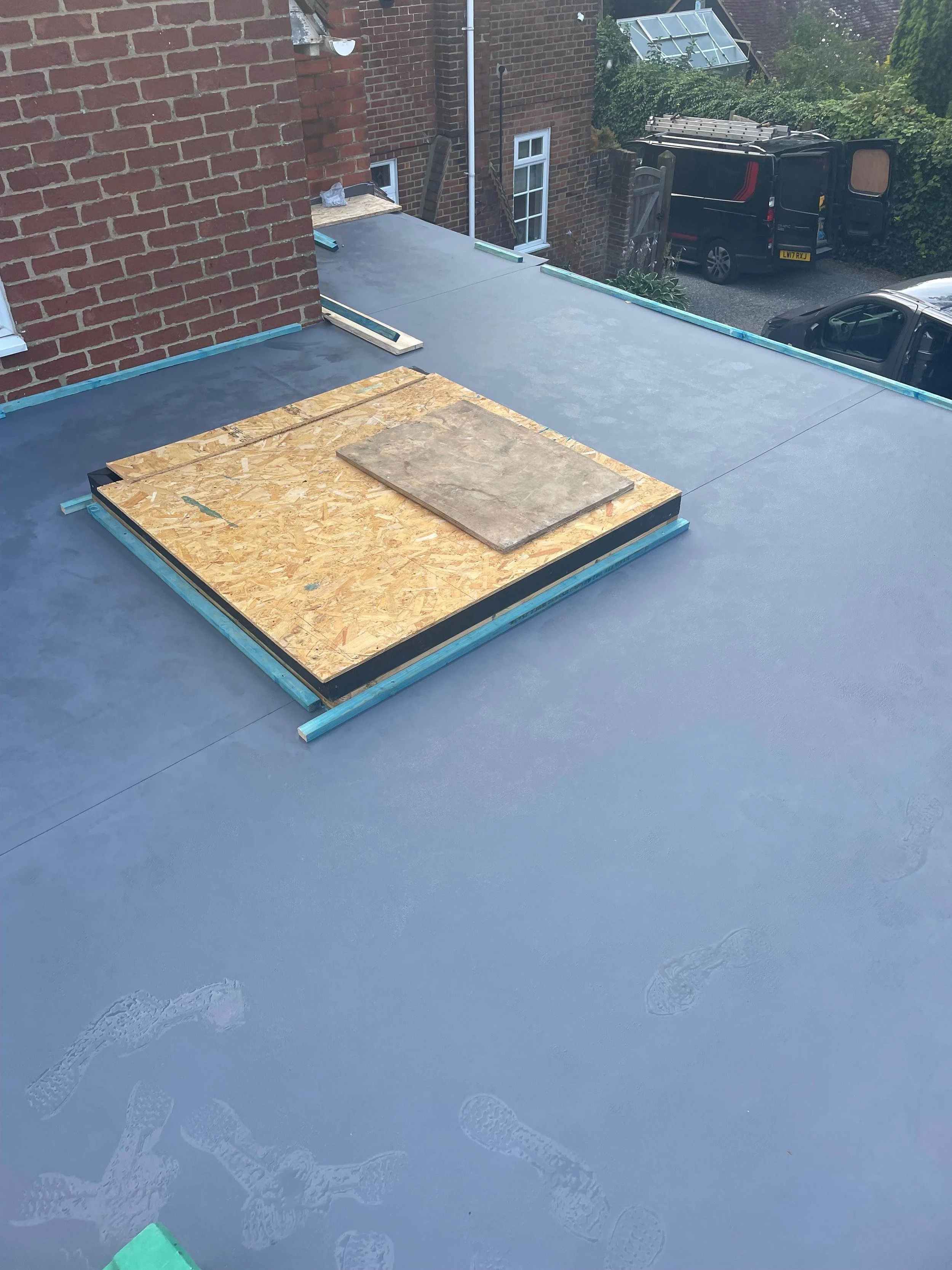 Flat roof under construction with a section covered with black waterproof membrane, nearby there is a larger area of new grey roofing material, and a small wooden platform with a piece of sheet on top. In the background, there are brick walls, a wind