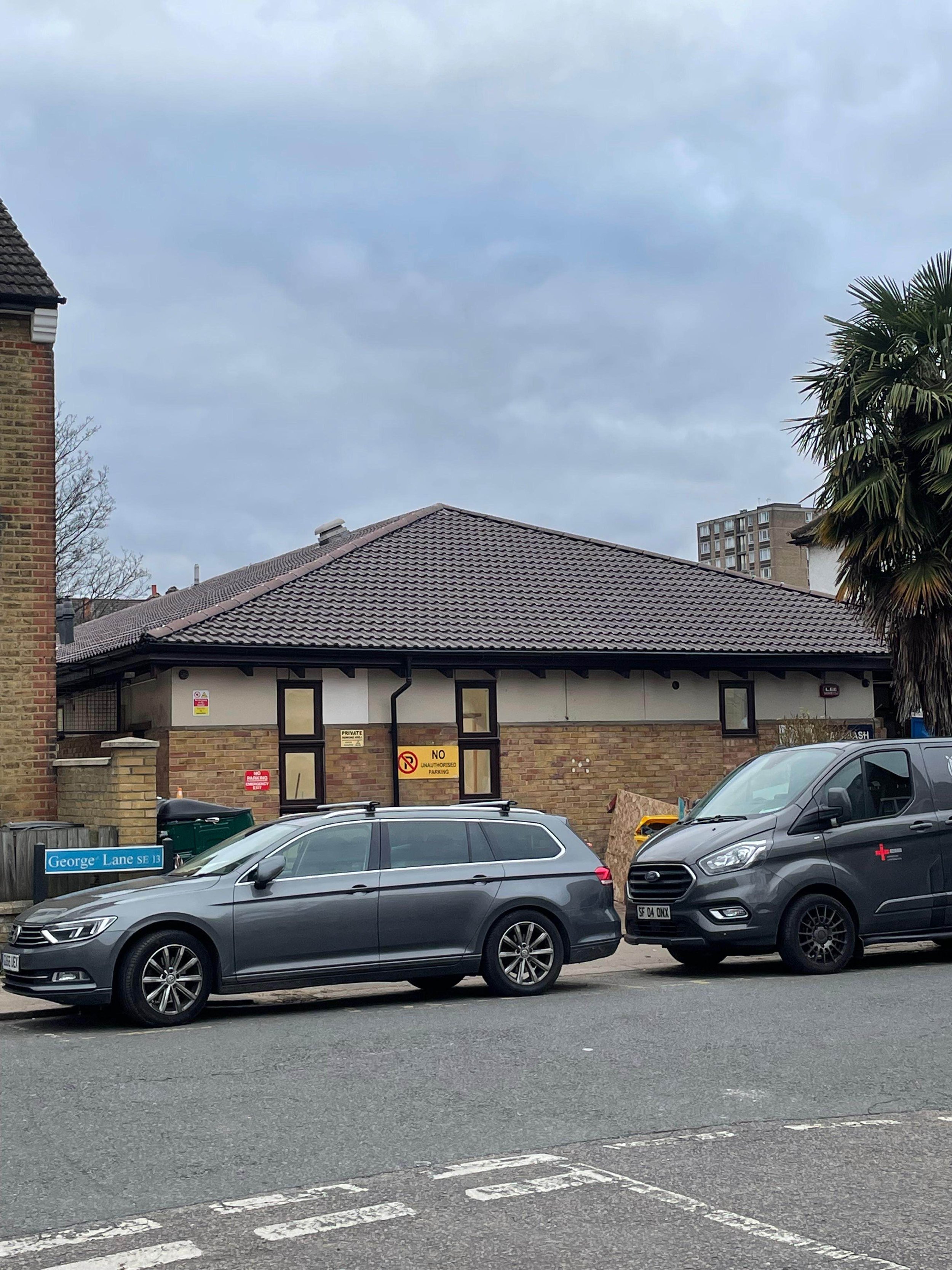 A street view showing two parked cars in front of a brick building under a cloudy sky. The car on the left is a silver station wagon and the one on the right is a black van with a red cross symbol.