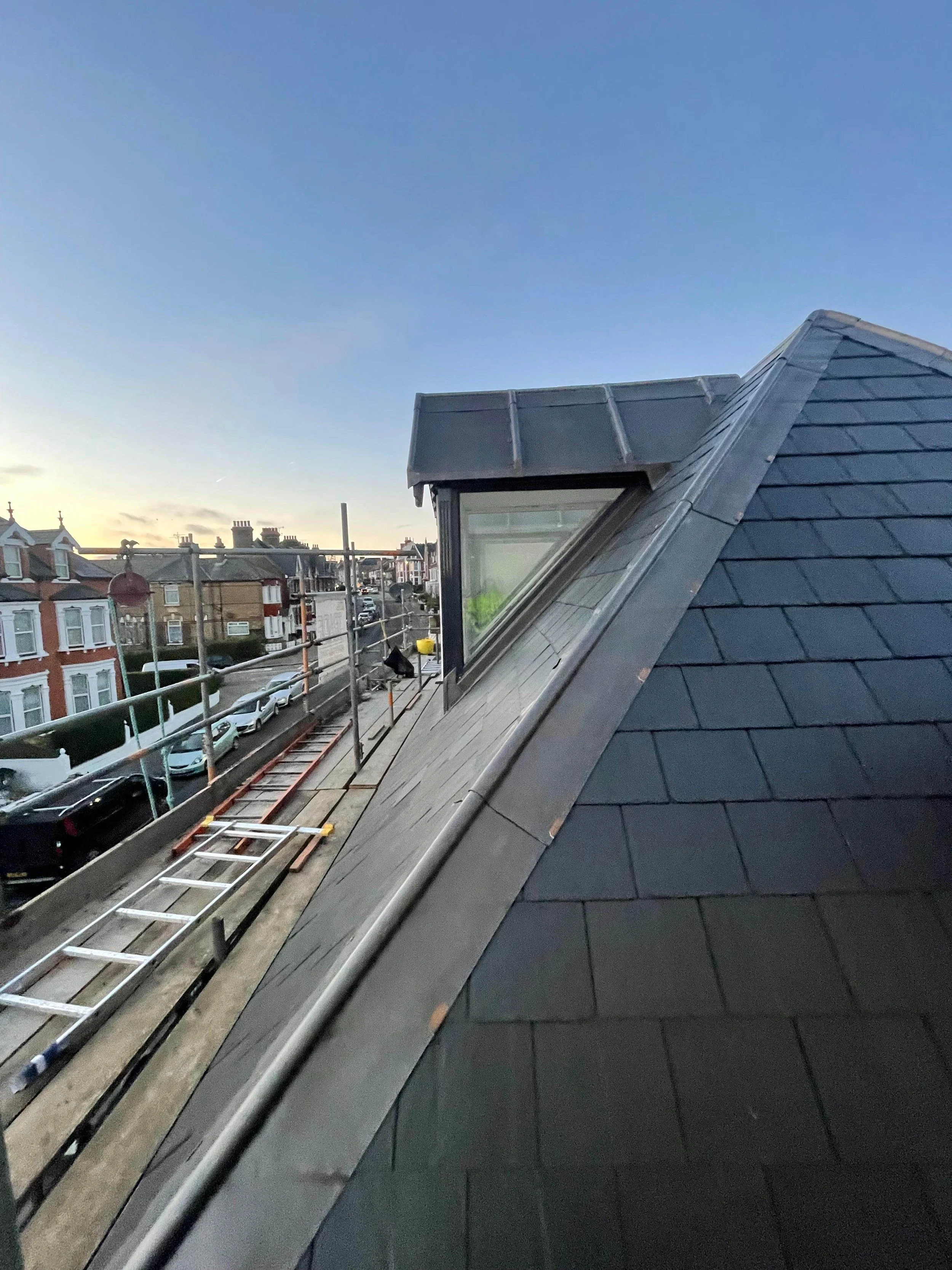 View from a rooftop construction site showing a sloped roof with dark shingles, a small dormer window, and a neighborhood with houses and cars in the background at sunset.