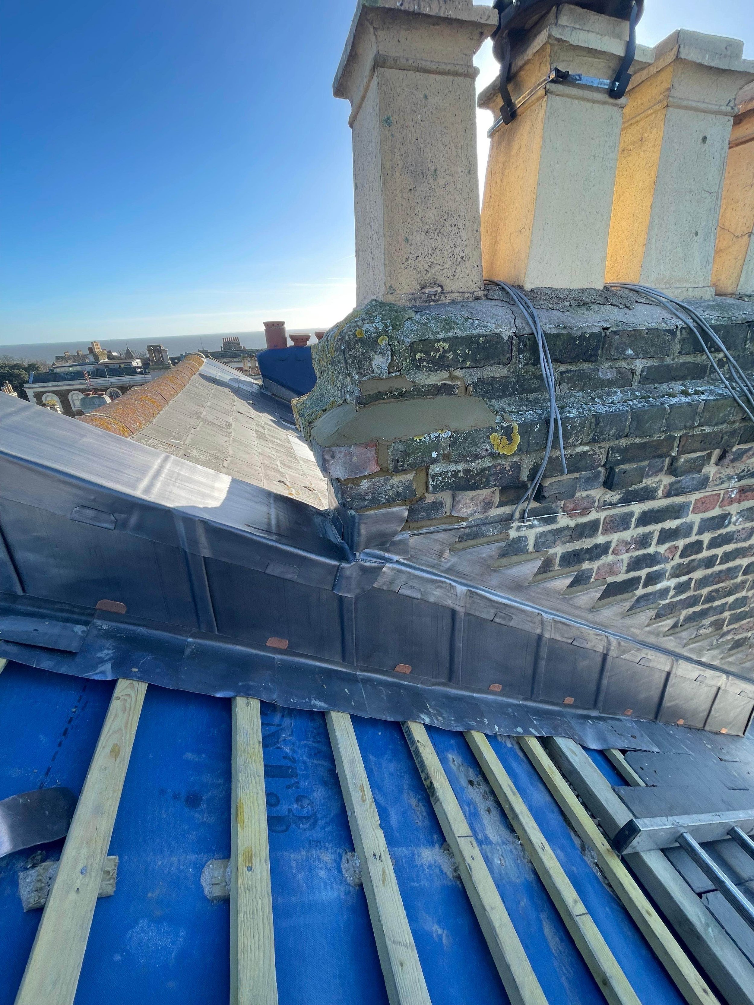 View of a rooftop with chimney stacks and building materials, including a metal flashing and partially constructed roof framework with blue underlayment and wooden battens, against a background of a cityscape and clear blue sky.