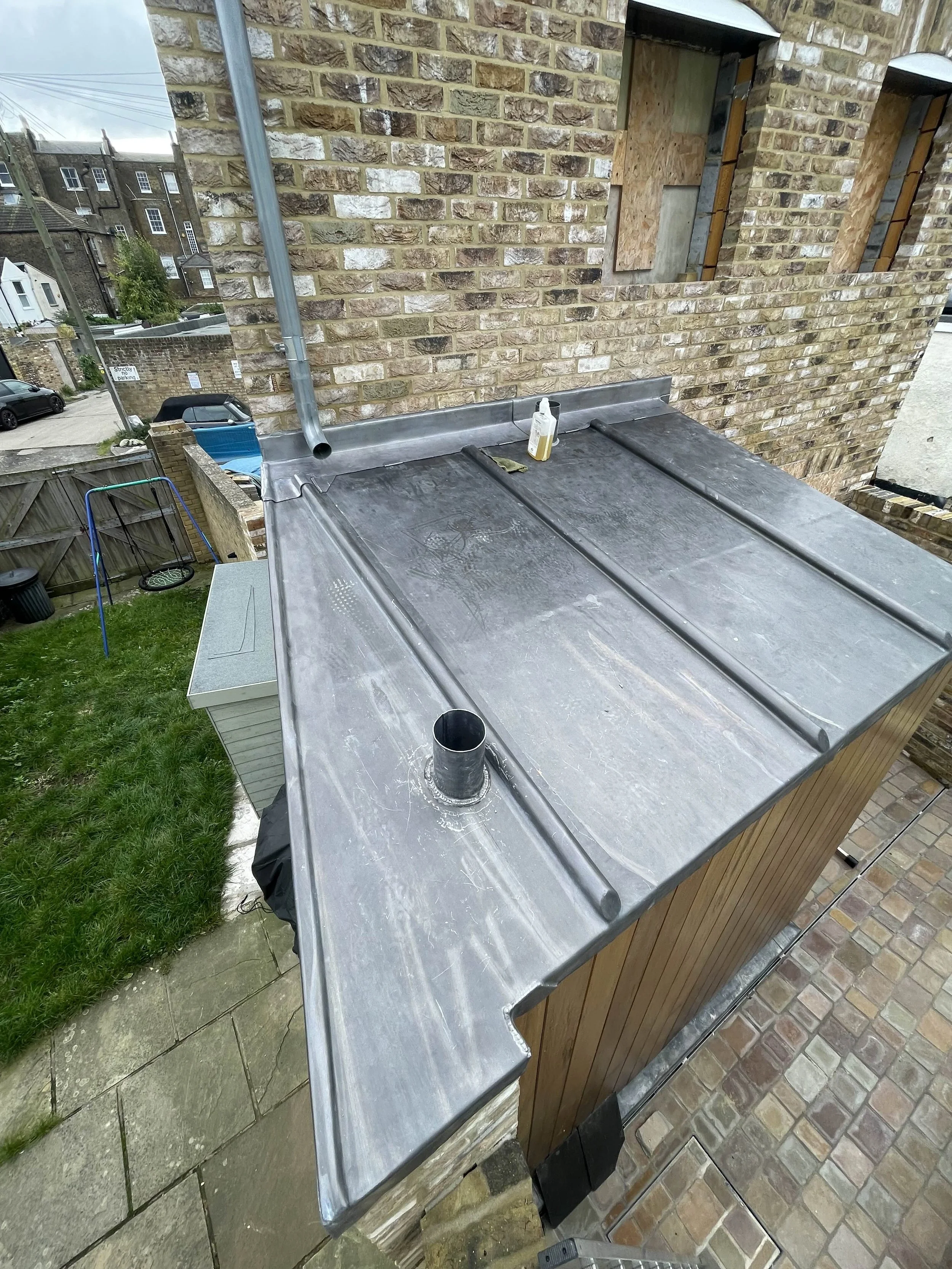 A metal roof extension on a brick building with a drain pipe, a white bottle, and a cloth. There's a grassy yard with a swing and a shed, and neighboring houses in the background.