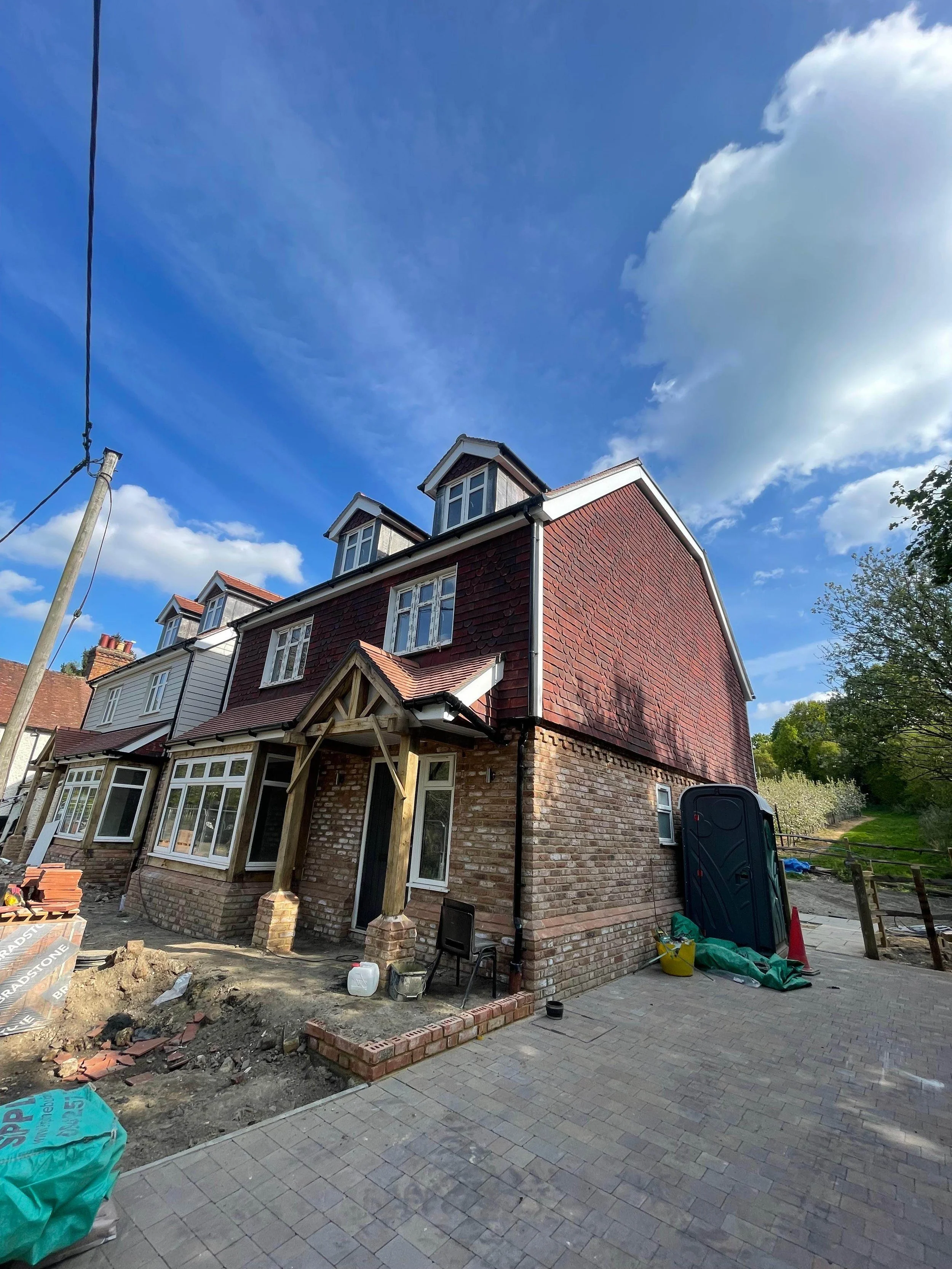 A brick house with a tiled roof under construction, with some scaffolding and building materials around, set against a bright blue sky with some clouds.