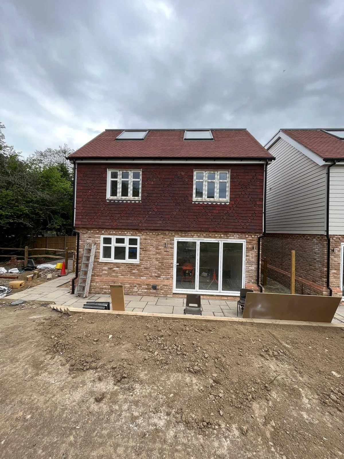 Under construction backyard with paving stones, construction materials, and tools in front of a two-story brick house with a red tiled roof, white-framed windows, and two skylights, with overcast sky.