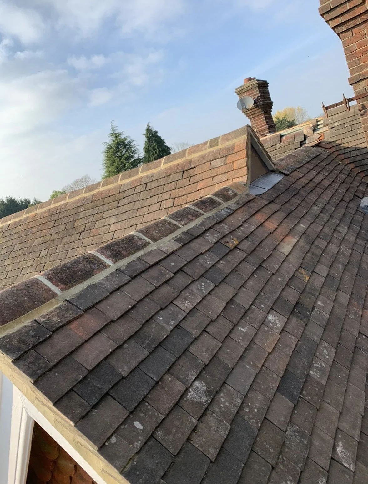 View of a sloped roof with red and brown shingles, brick chimneys, and a partly cloudy sky in the background.