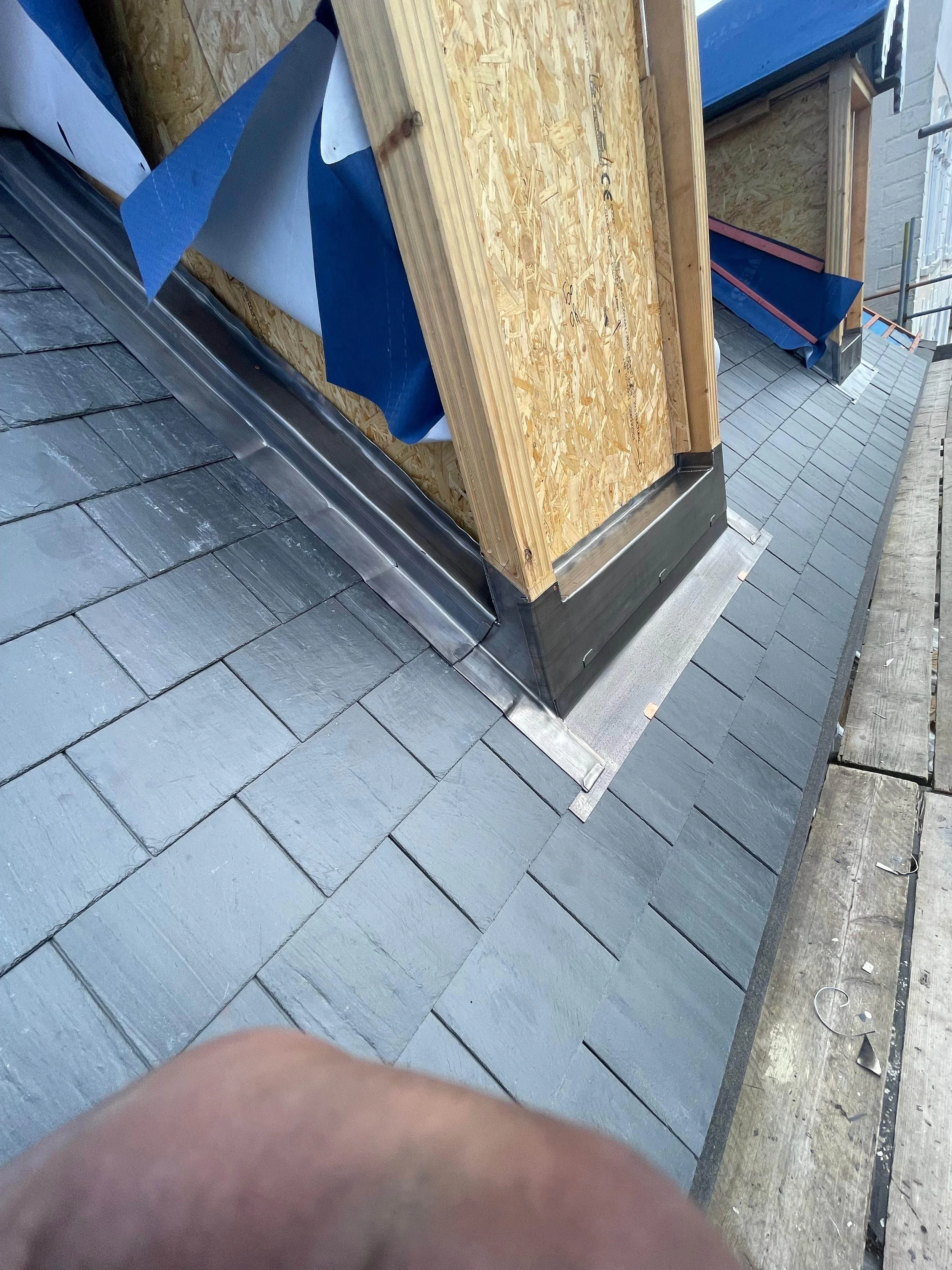Construction site showing a roof with gray shingles, a wooden structure with a diagonal metal flashing, and blue and white roofing underlayment in the background.