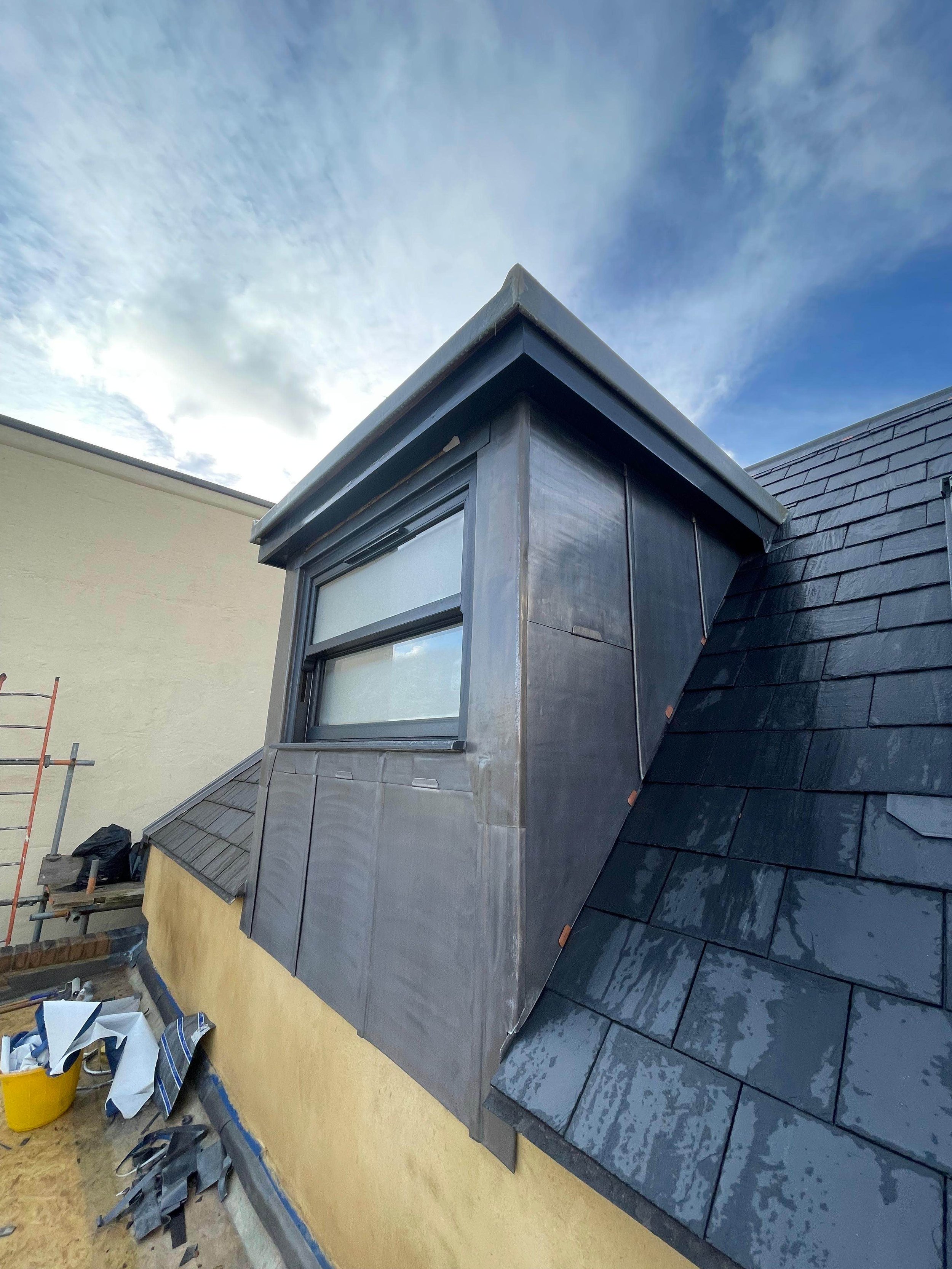 Close-up view of a building's rooftop with black shingles and a window, under a cloudy sky.