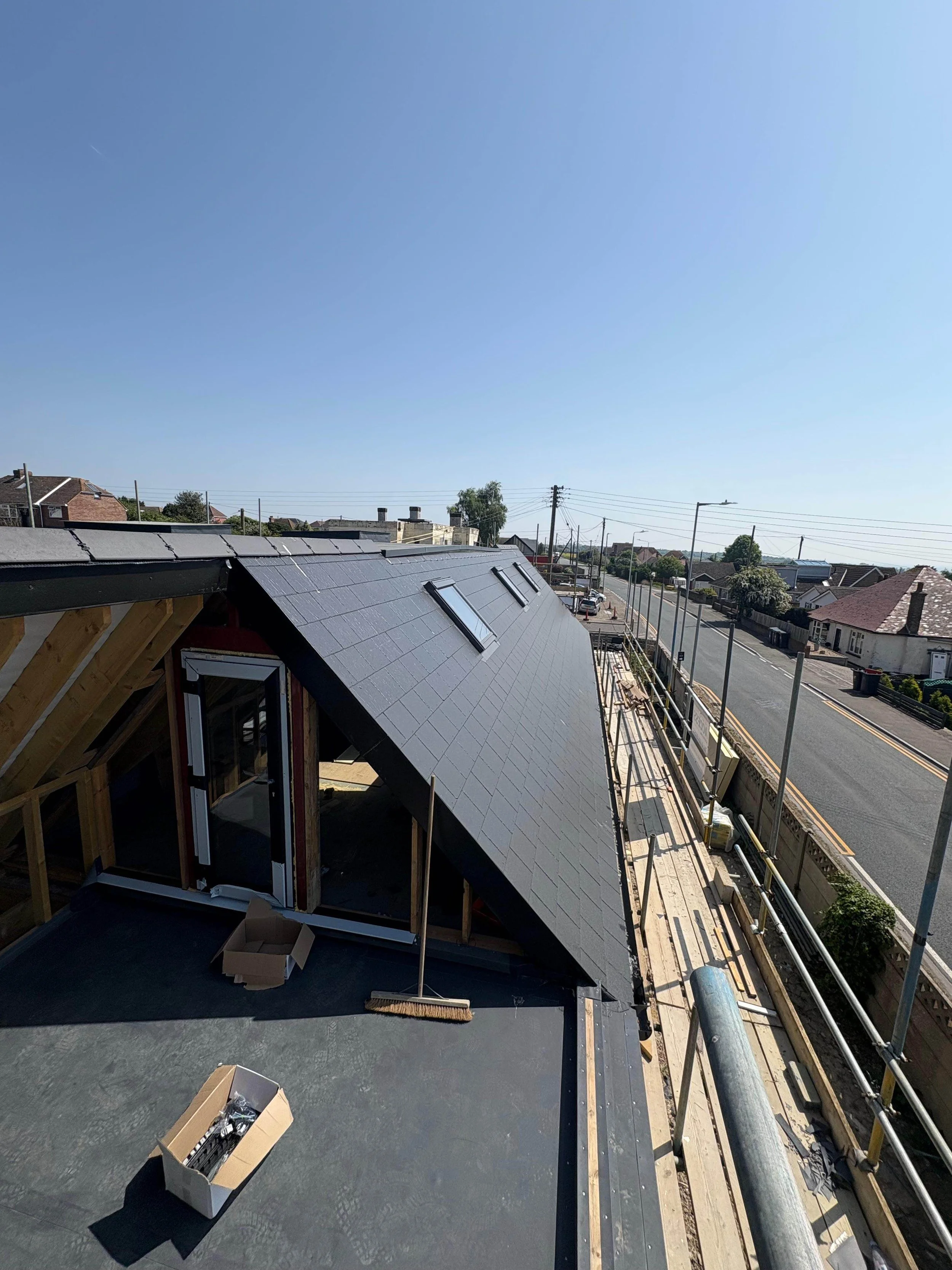 Upward view of a house under construction, with a new black tiled roof and a partially finished structure, scaffolding along the side, and a suburban street in the background.