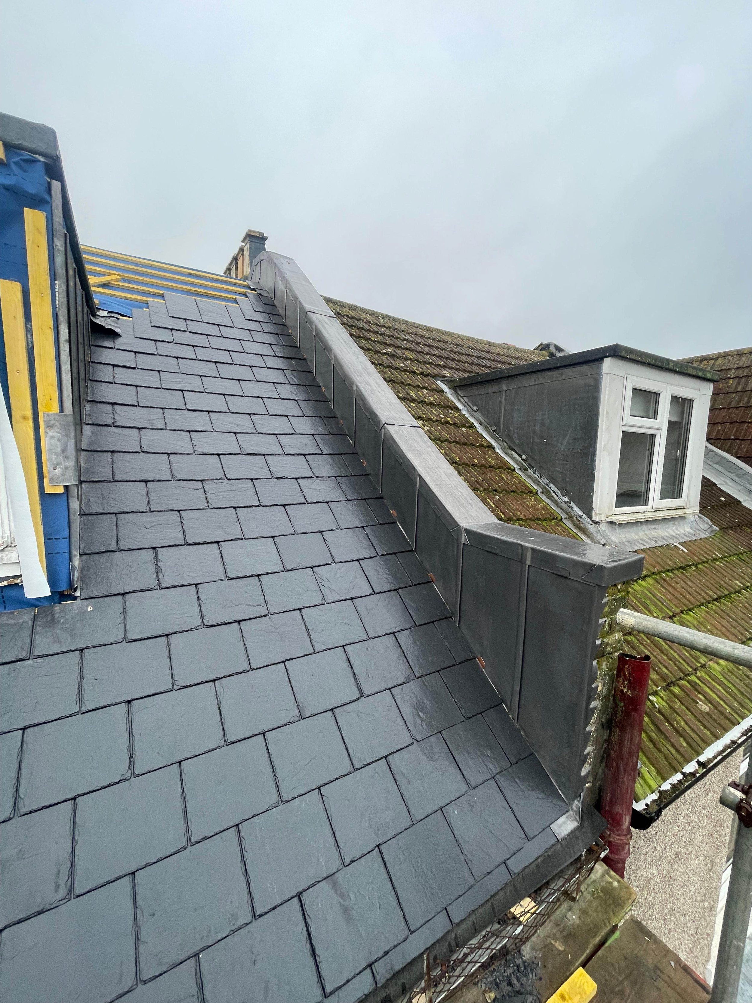 Roof of a building with new dark gray slate shingles and a chimney with roofing material. Adjacent roof with moss and weathered tiles, with a small dormer window.