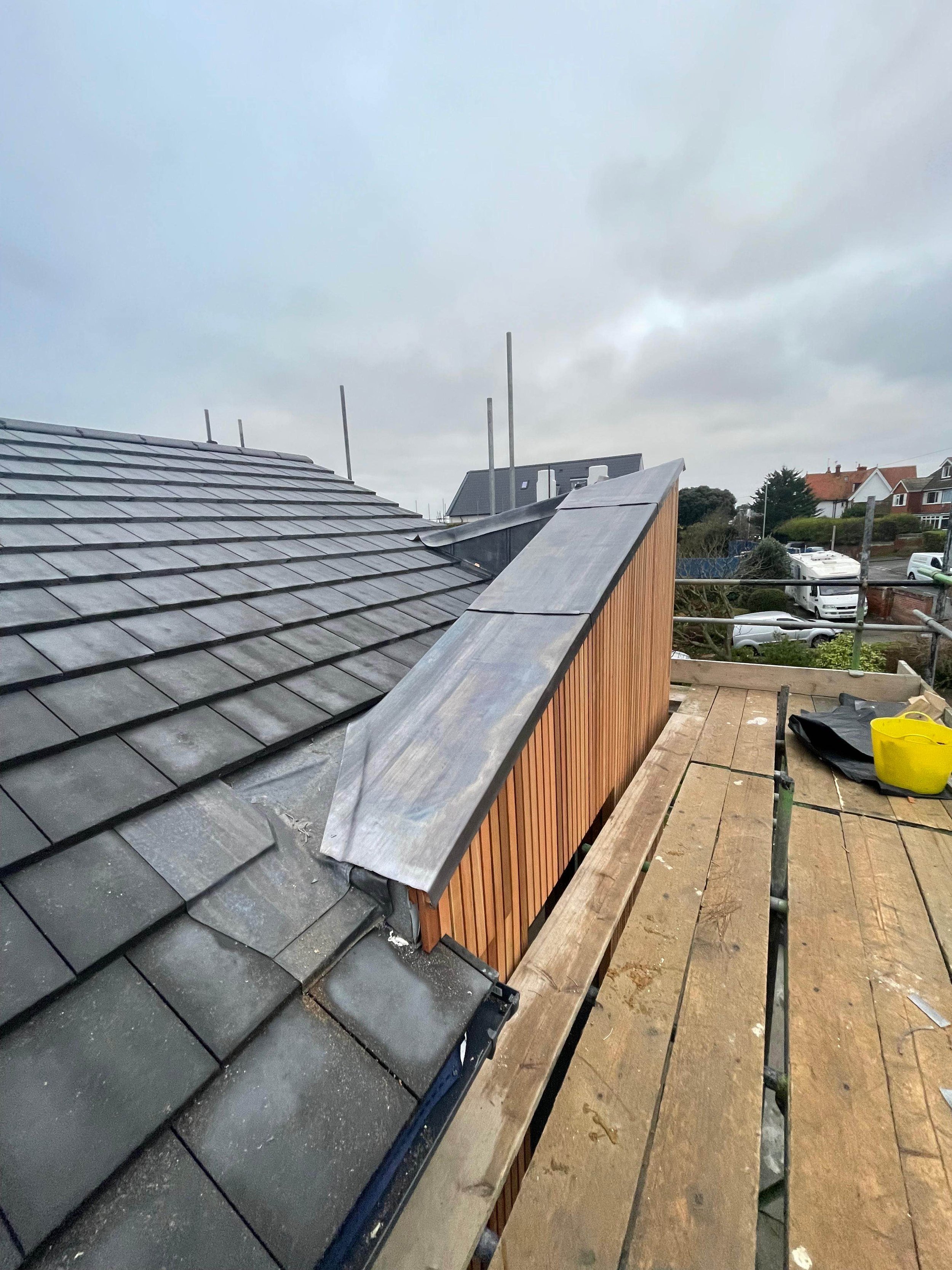 Rooftop under construction with gray tiles, metallic flashing, wooden siding, and scaffolding, overlooking a suburban neighborhood with houses and cars.