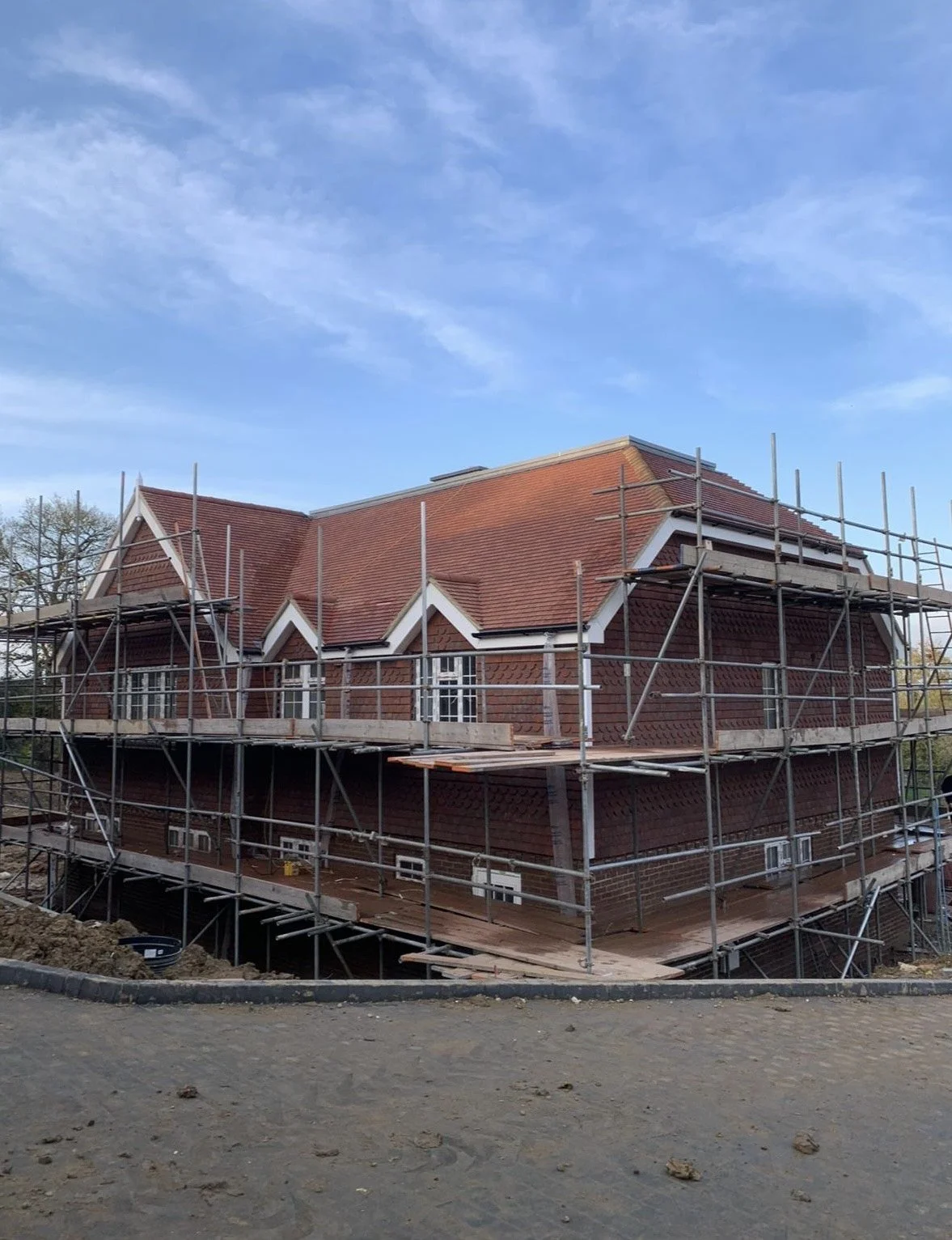 A house under construction. The house has a red tiled roof and brick walls. Scaffolding surrounds the building, and the sky is blue with some clouds.