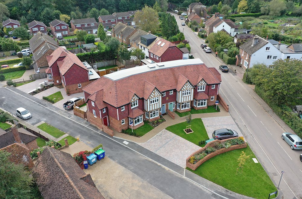 Aerial view of a residential neighborhood with a large, modern red brick building, multiple houses, cars parked along the roads, green lawns, and trees.