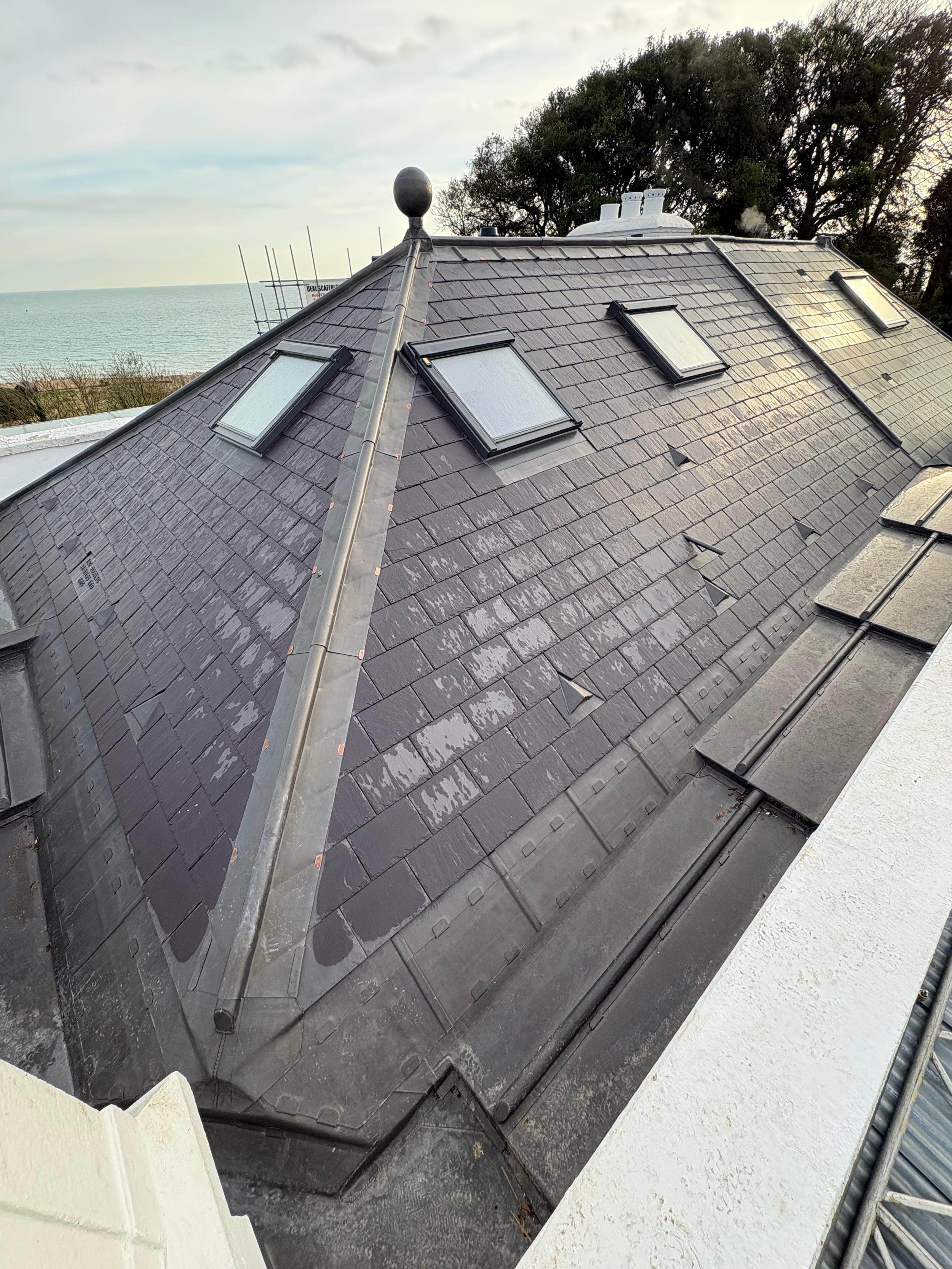 View of a rooftop with dark slate shingles, skylights, and a metal ridge cap, overlooking the ocean and trees in the background.