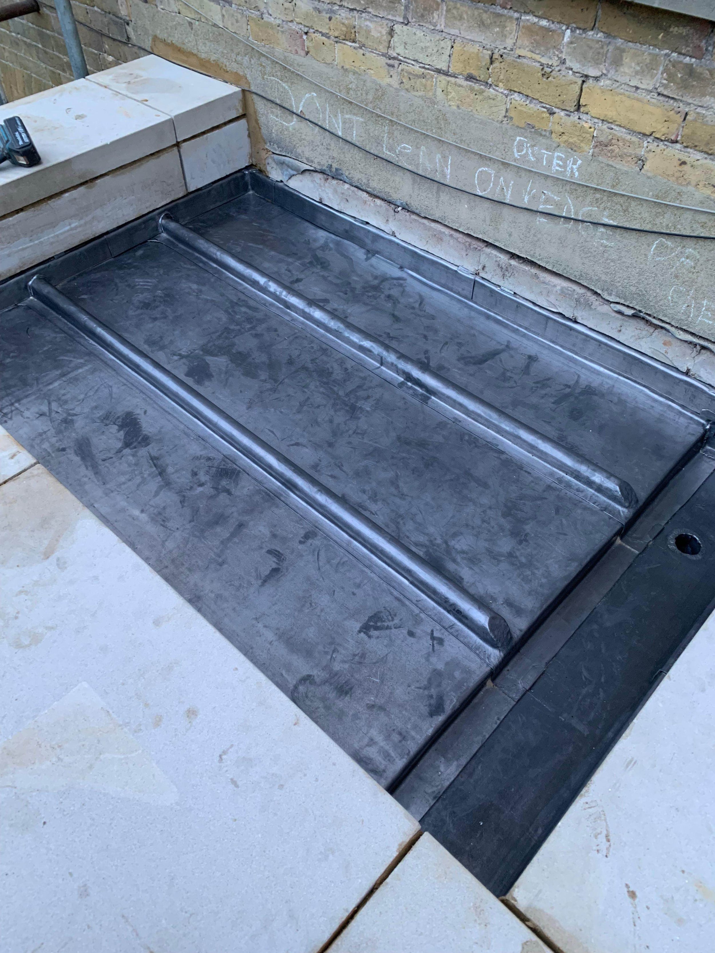 Metal firepit or water feature frame partially installed on a brick patio with surrounding brick wall and beige tiles.