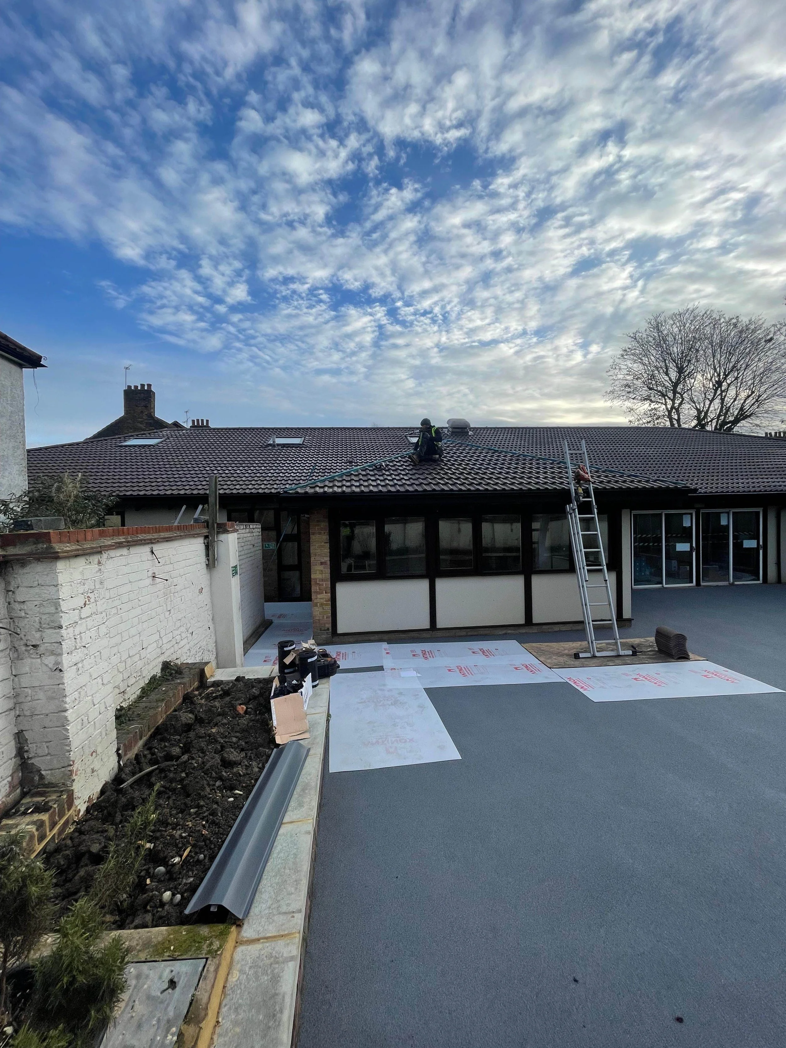 Construction worker on roof installing tiles with ladder and tools nearby, clear sky with clouds, building with large windows and parking area in front.