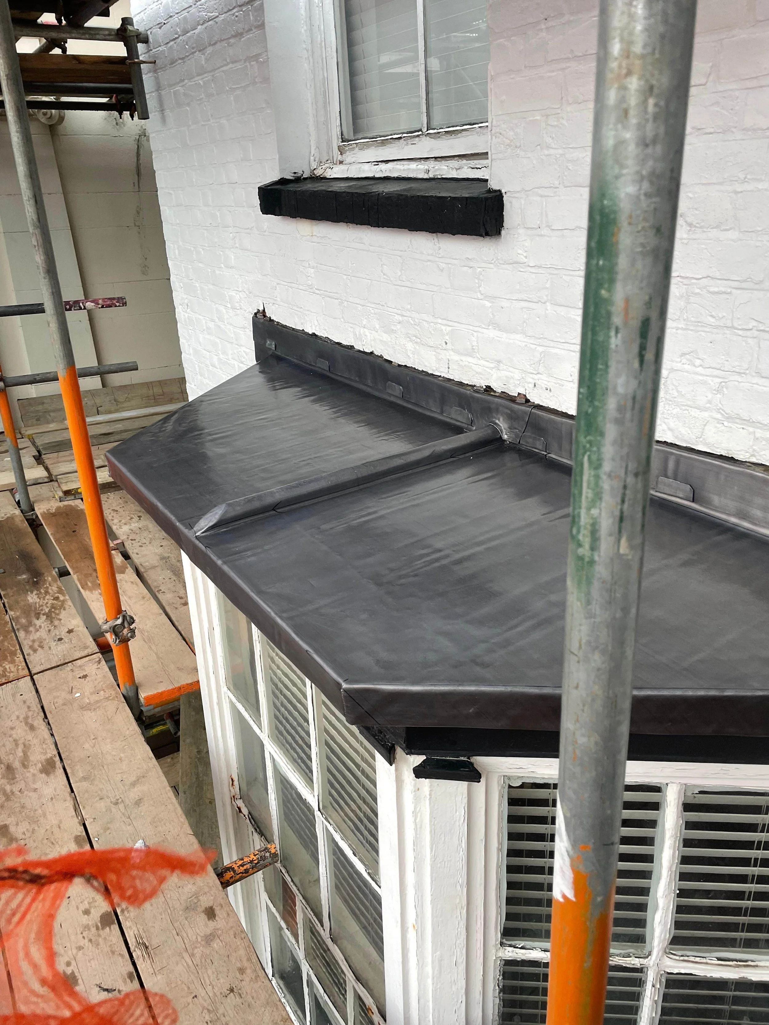 A black metal awning installed above a bay window on the exterior of a white brick building, with scaffolding around it during construction or repair.