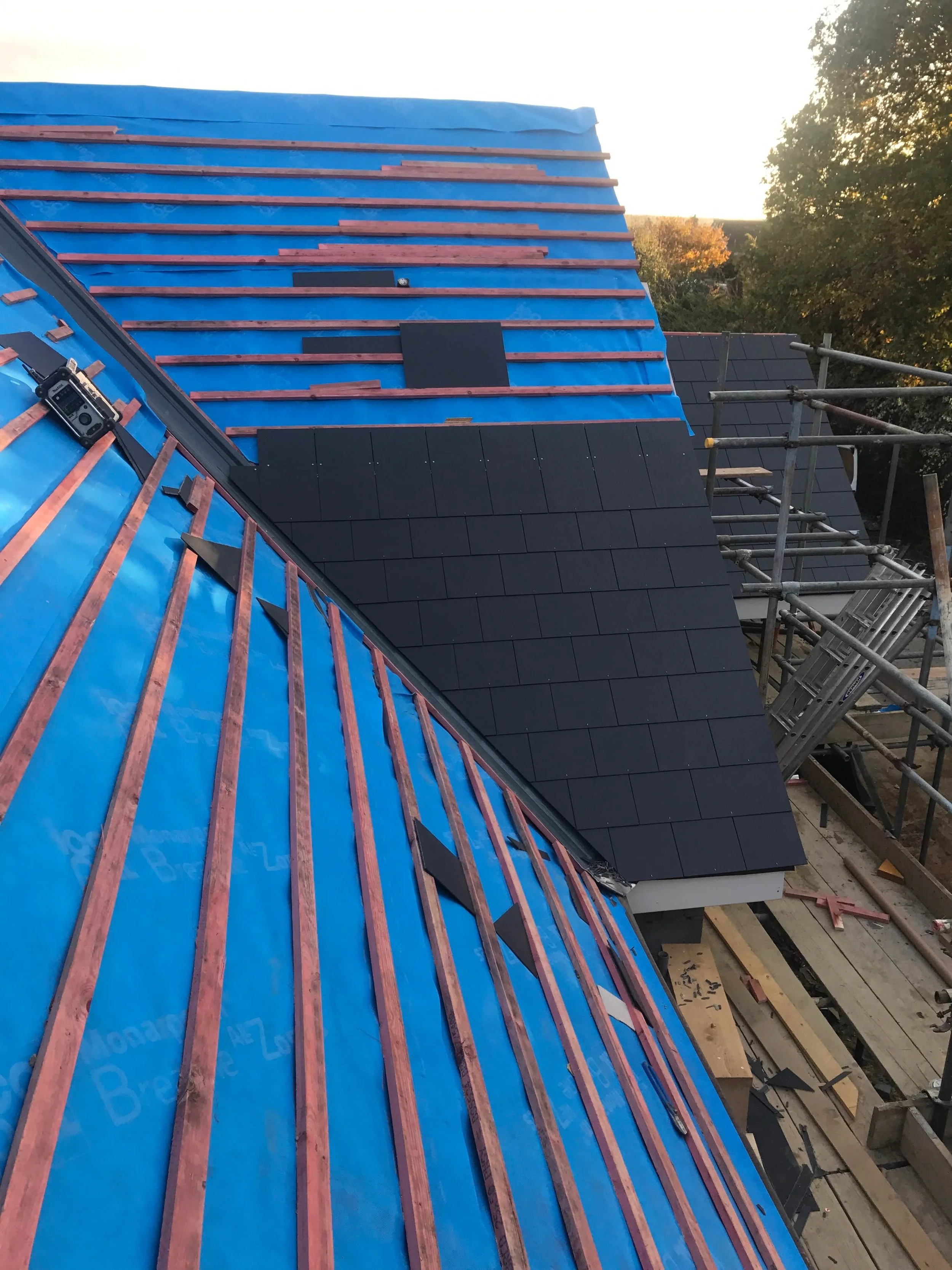Construction site showing a roof with blue underlayment and copper roofing strips, partly covered with dark shingles, scaffolding on the side with trees and sky in the background.