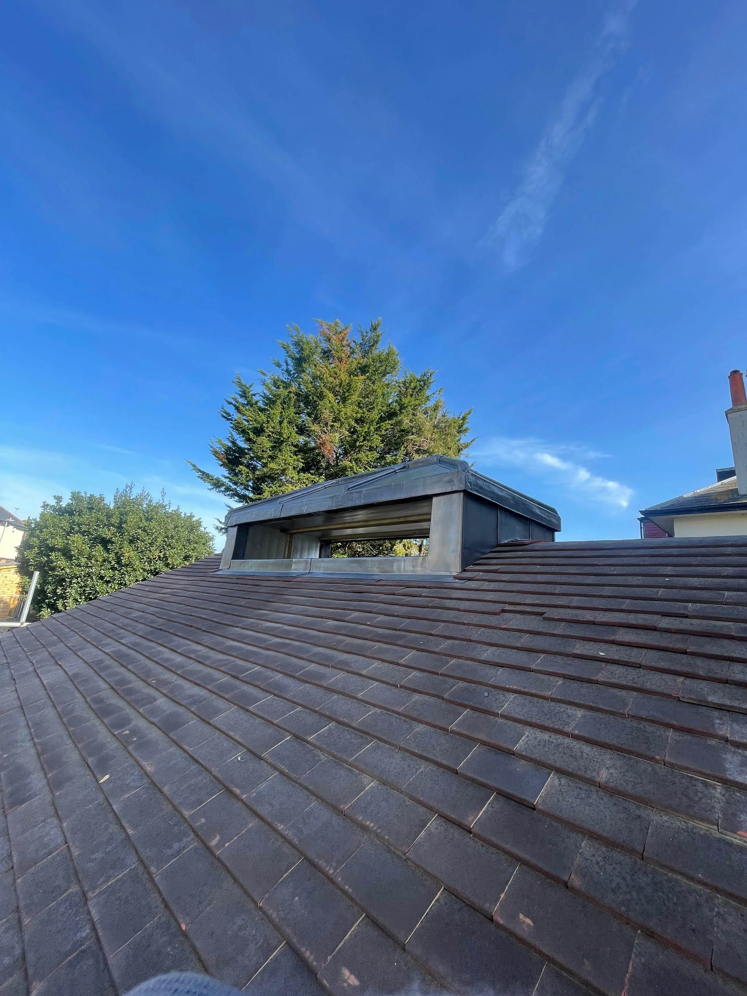 View of a roof with asphalt shingles, a metal chimney cap, a tree in the background, and a blue sky with some clouds.