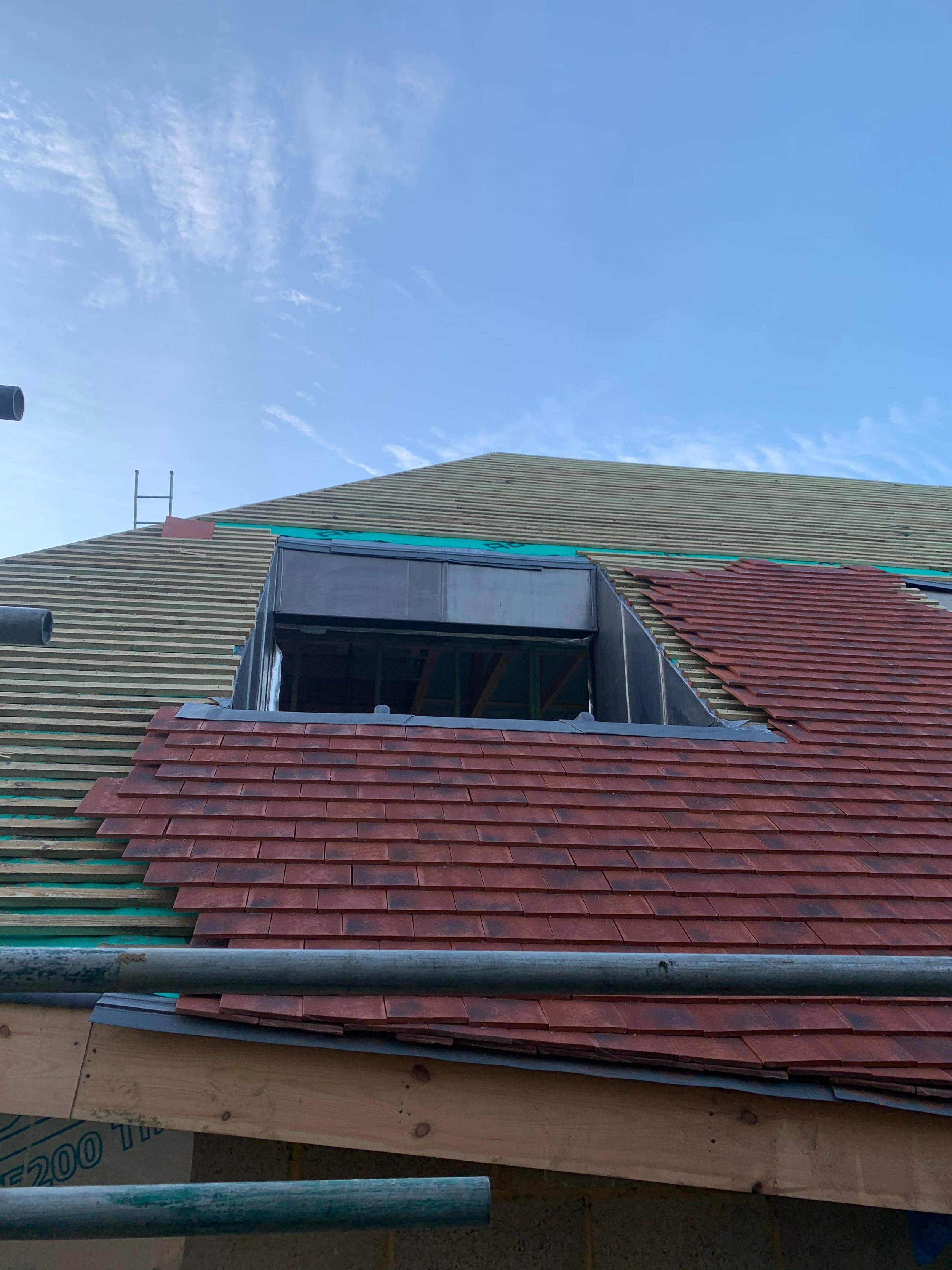 A house under construction with a partially completed roof, showing new red roof shingles and an open space where a dormer window will be installed, against a blue sky with a few clouds.