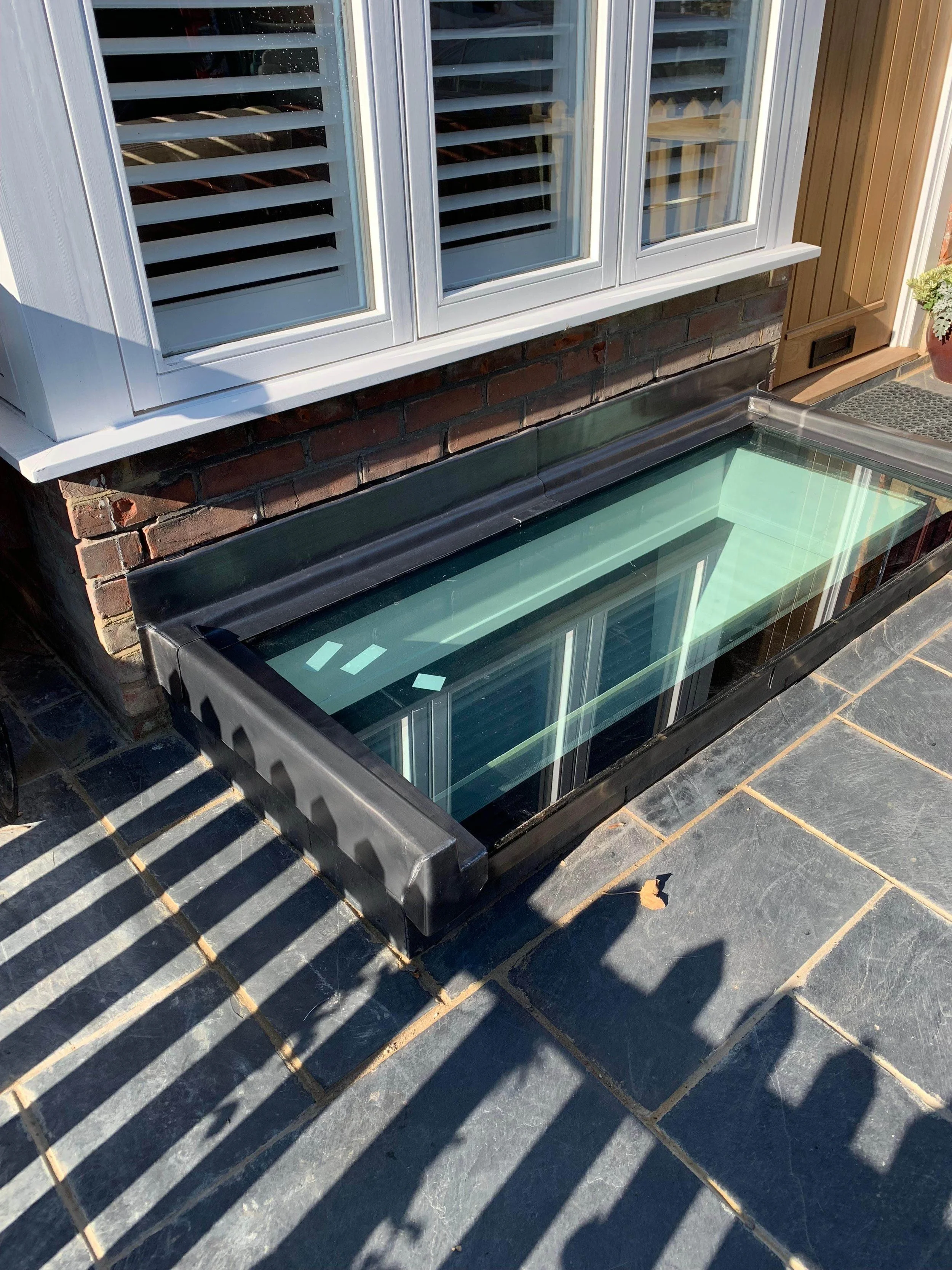 A glass window well for a basement or cellar, installed outside a house with brick and wood exterior, on a stone-paved ground.