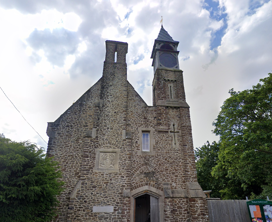 An old stone church with a tall steeple, surrounded by green trees, under a partly cloudy sky.
