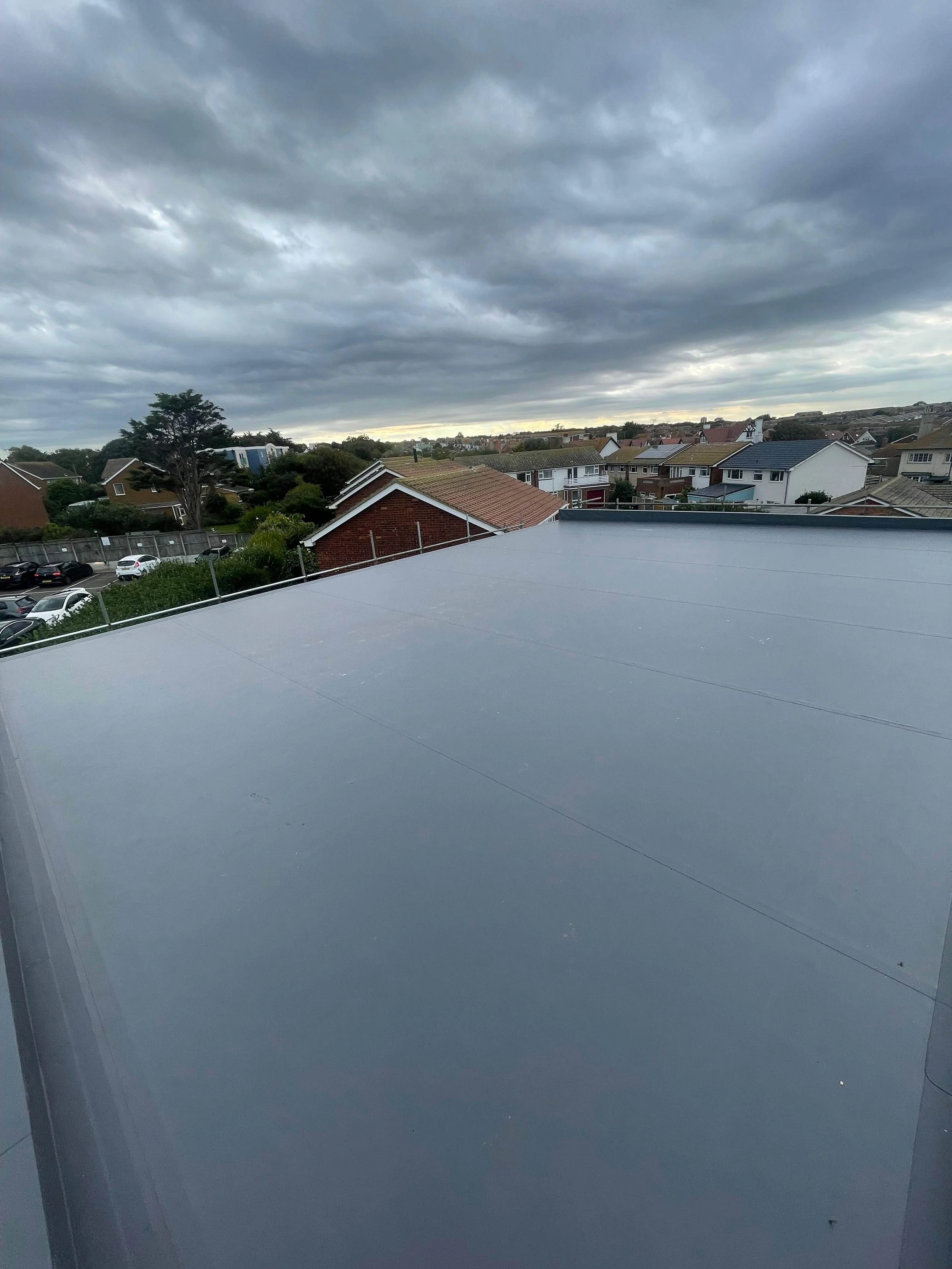 View of a cloudy sky over a residential neighborhood with houses, trees, and parked cars, as seen from a rooftop with a metallic surface.