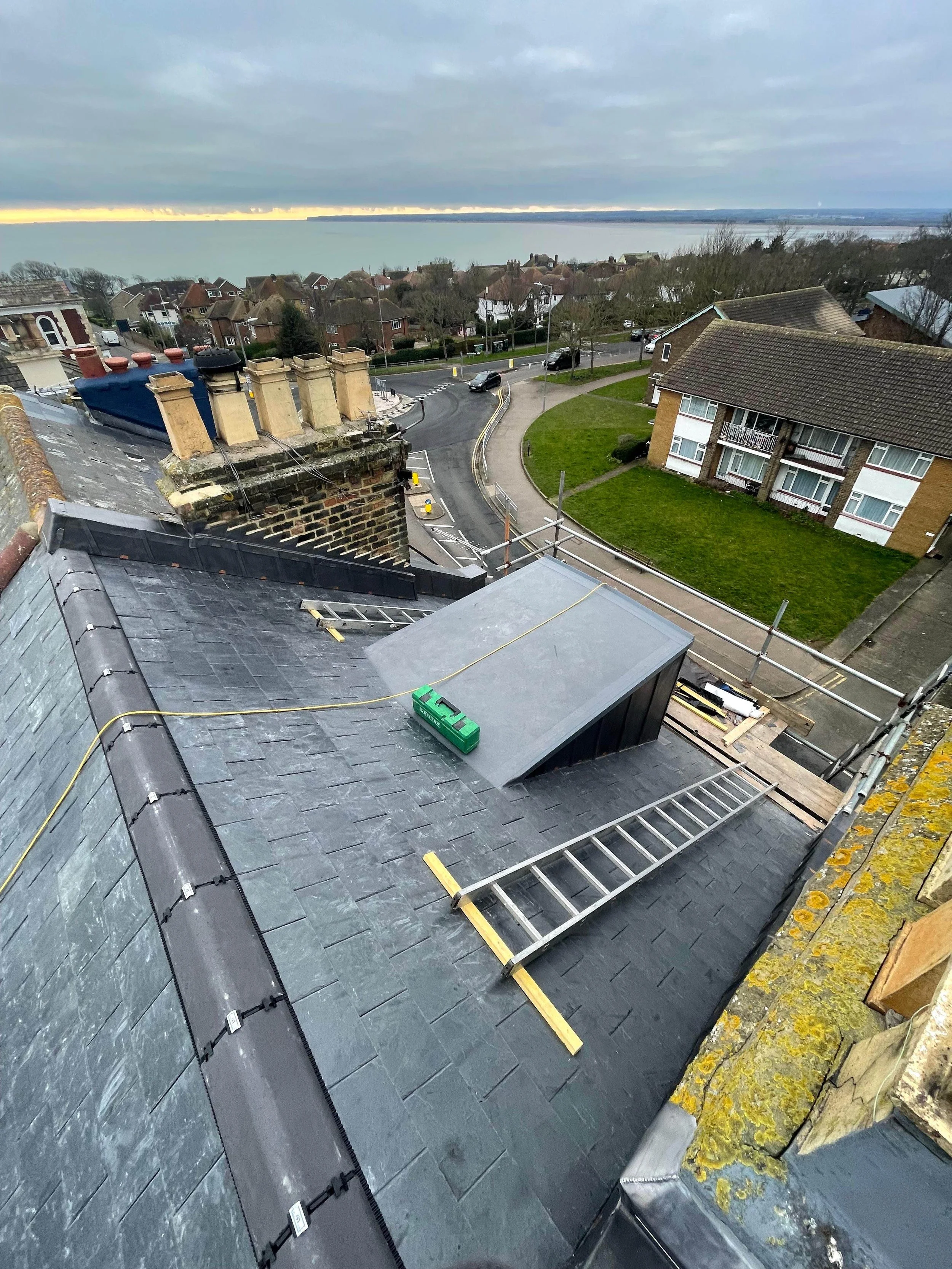 View from a rooftop showing construction or renovation work, including a ladder, electrical tools, and a small structure on the roof. The background features a residential neighborhood with houses, a parking road, and a body of water under a cloudy s