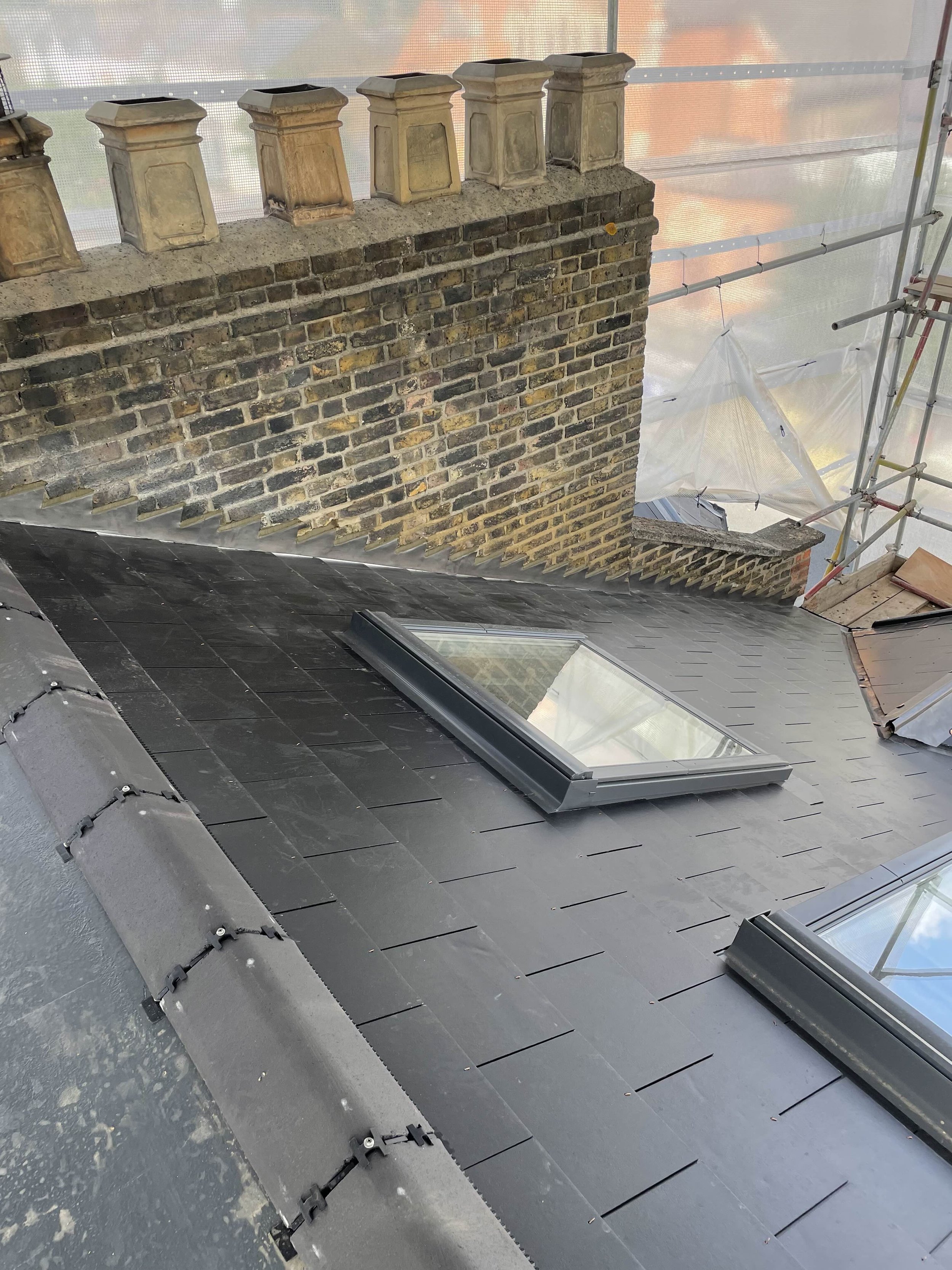 View of a roof with black tiles, roofing skylights, and an old brick chimney with decorative chimney pots, with scaffolding and building materials nearby.