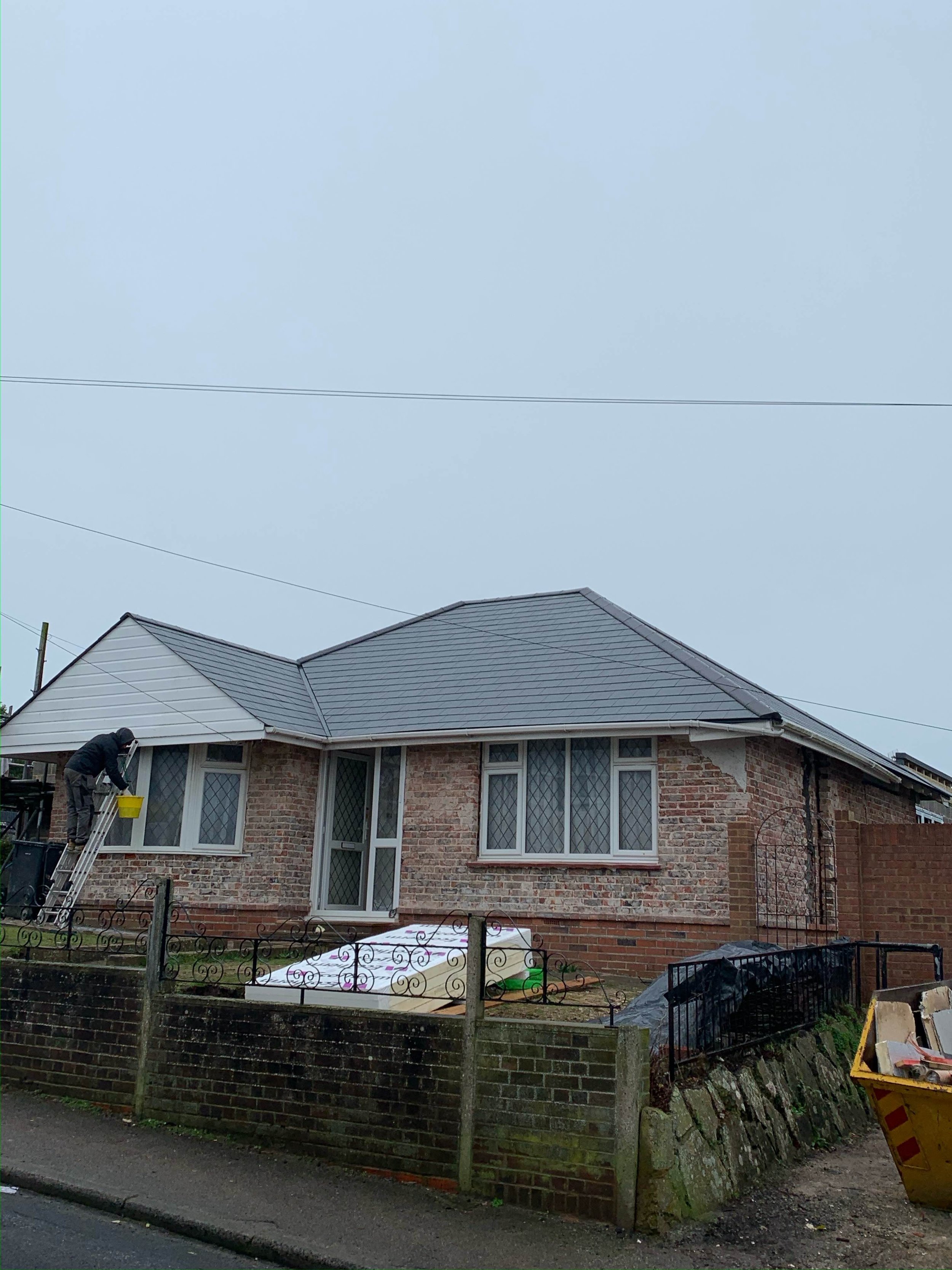 A person cleaning windows on the side of a brick house with a ladder, in a residential neighborhood.
