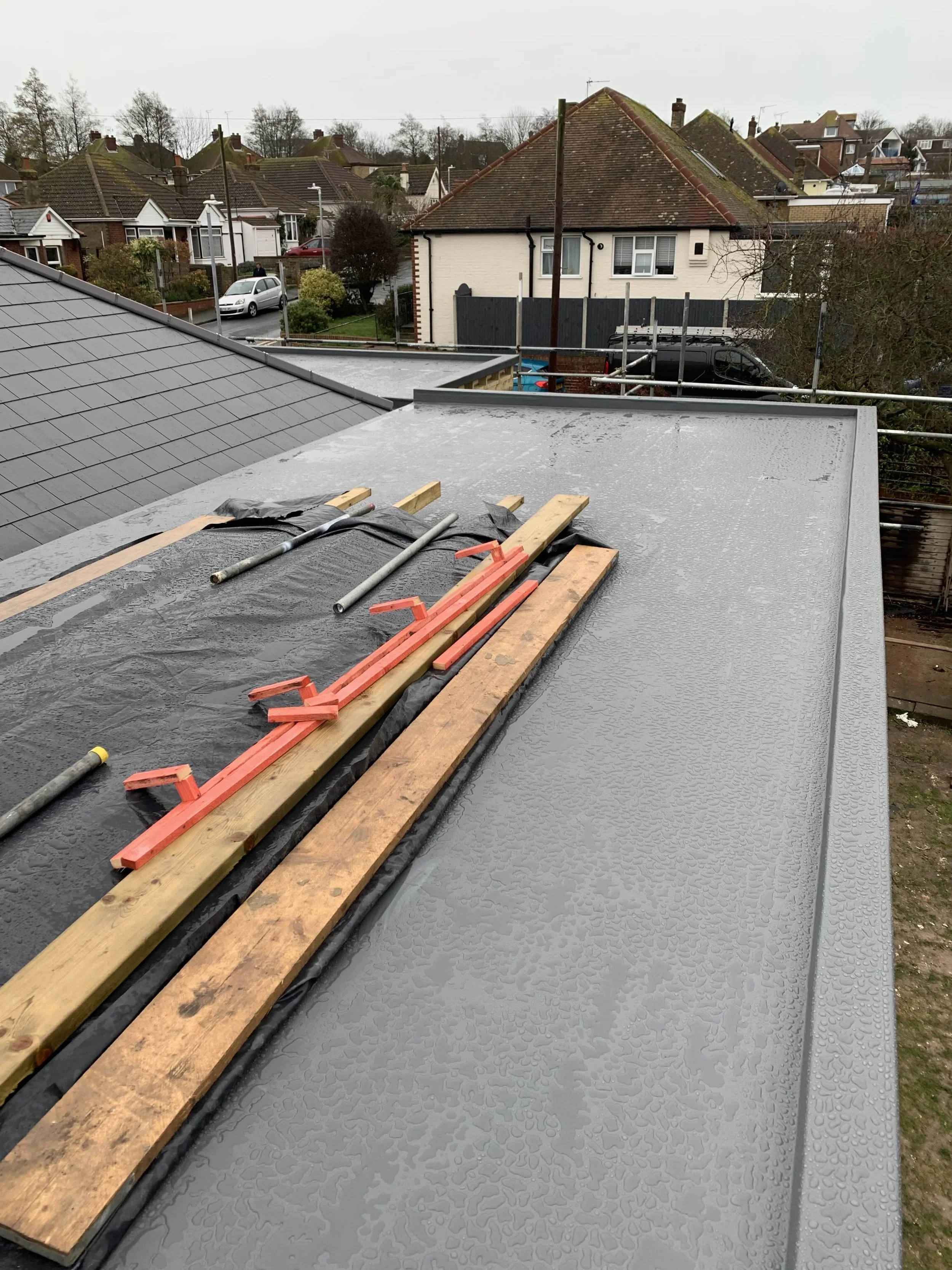 A flat rooftop under construction with wooden and metal tools, covered partially with black material, and another section with a gray textured surface. Residential houses and trees are visible in the background.
