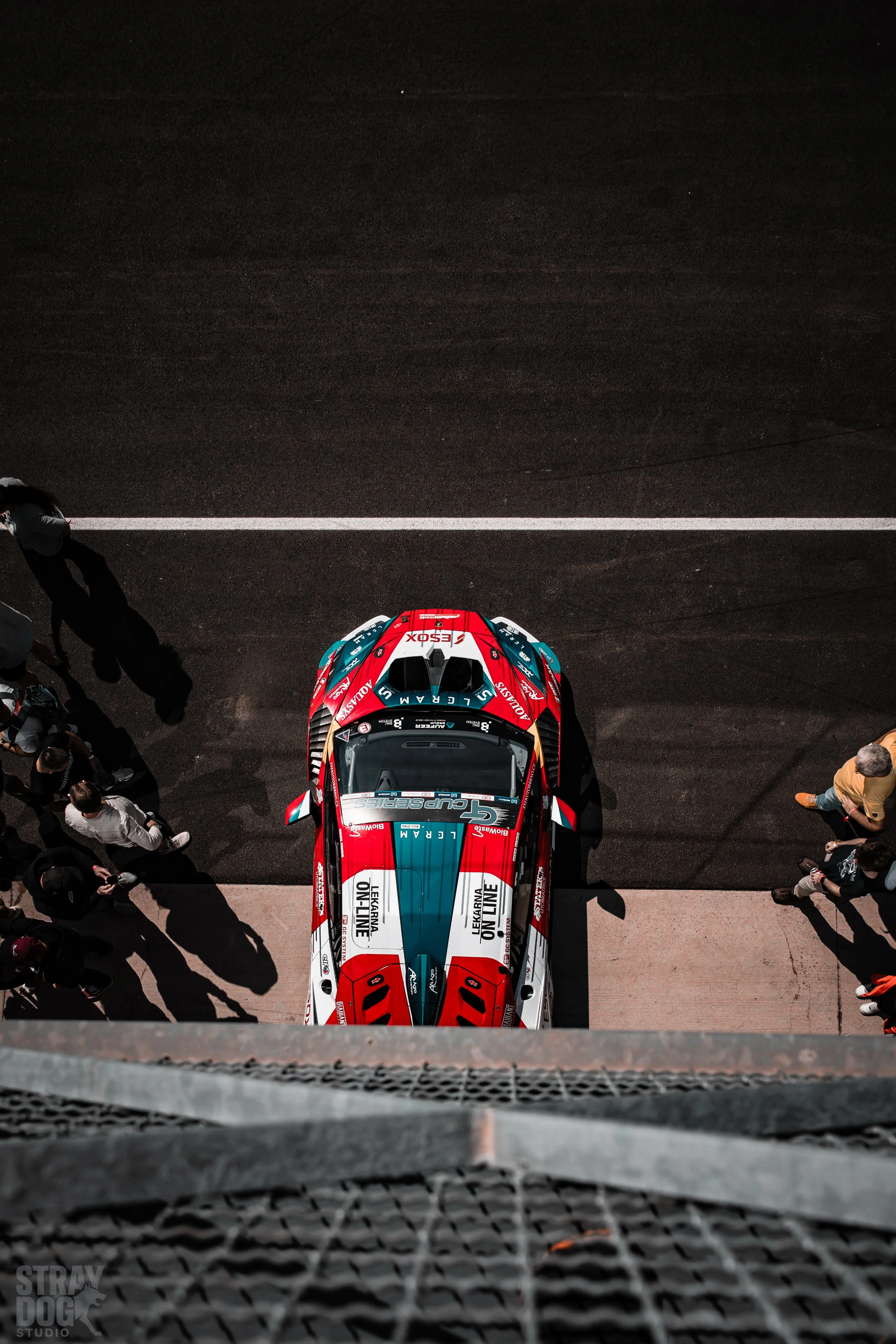 Top-down view of a race car parked in pit lane with spectators around, taken from an elevated position.