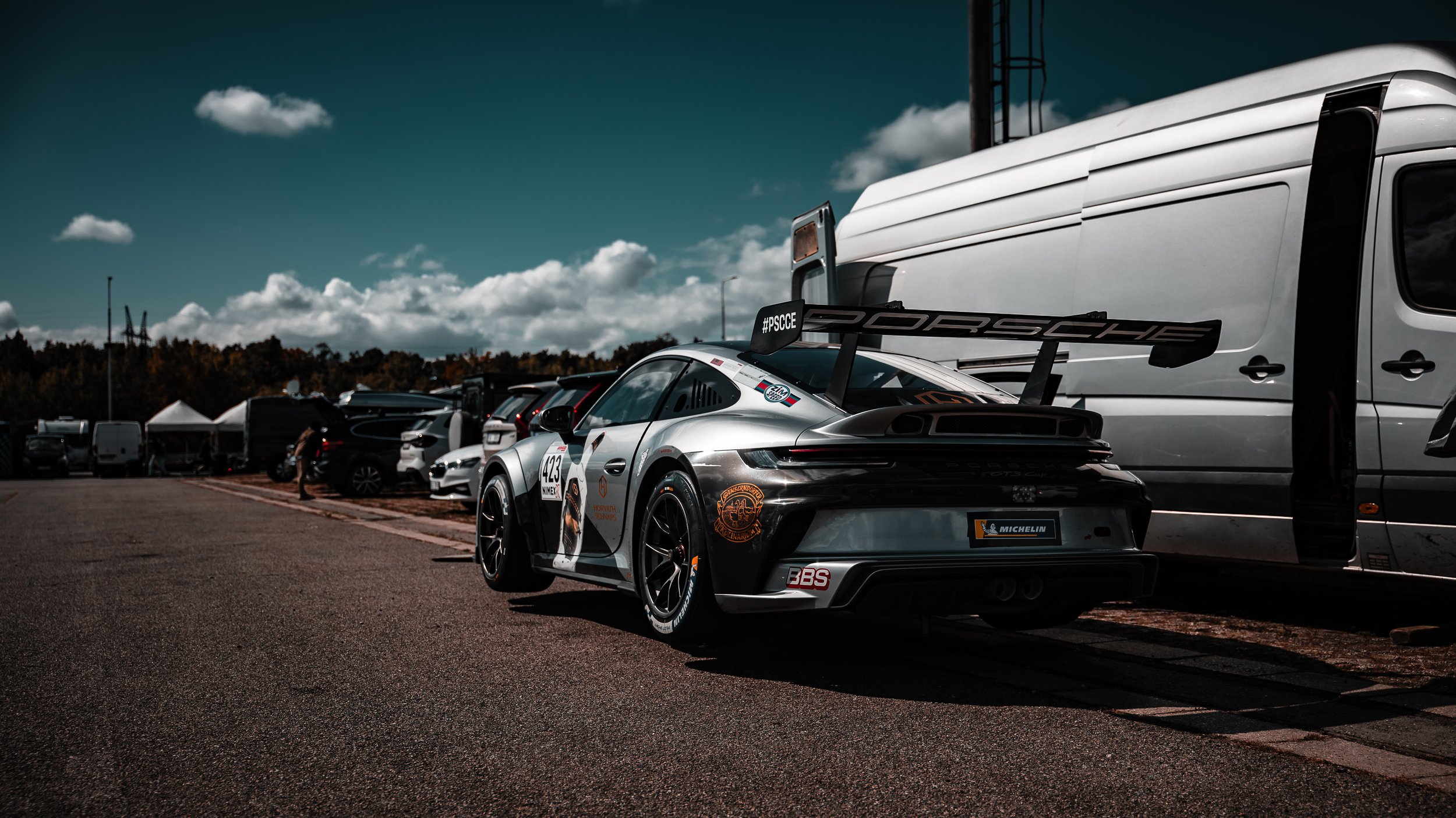 Silver Porsche race car with rear spoiler parked in a paddock lane, surrounded by other vehicles and tents under a cloudy sky.